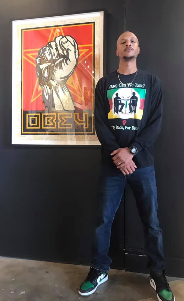Young man stands with arms crossed in front of a framed artwork of a raised fist on a red background.