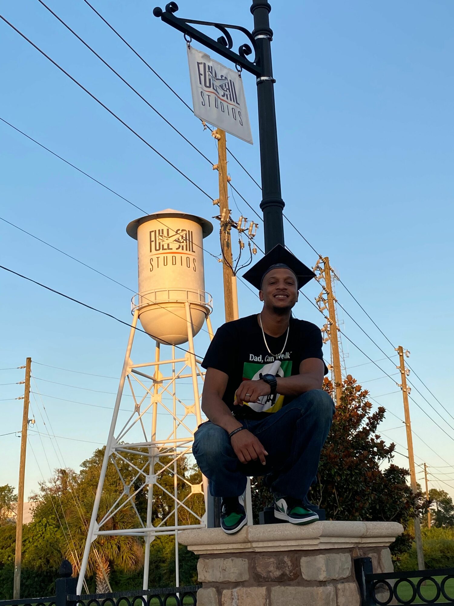Young man sitting on a stone ledge outdoors, smiling, wearing a cap and casual clothes, with a water tower and power lines in background.