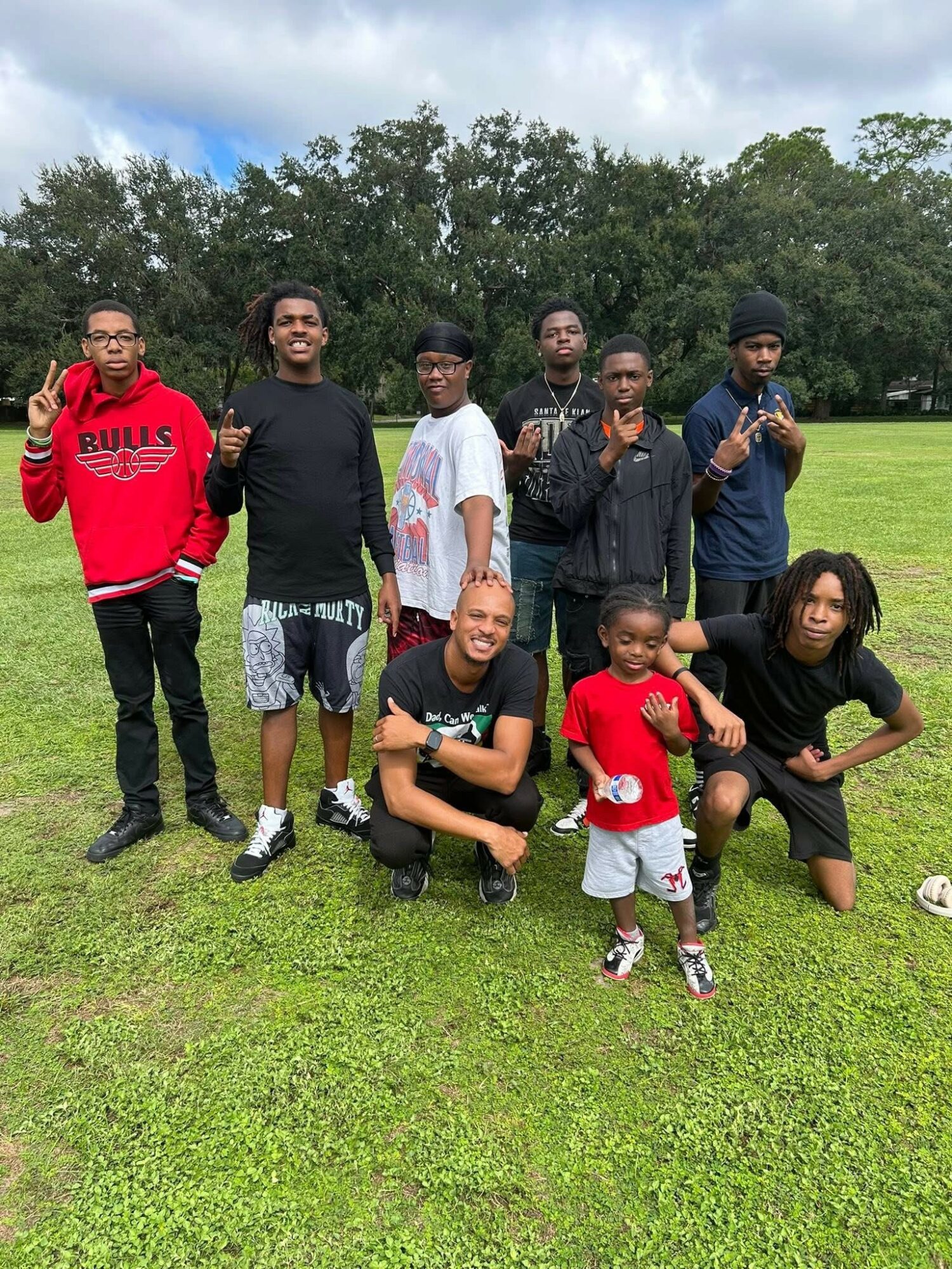 Group of nine people, including children and adults, standing and kneeling on grass in a park with trees and cloudy sky.