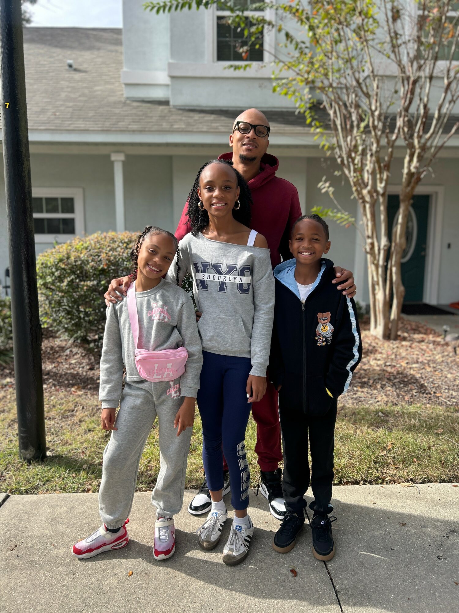 Four people standing outdoors in front of a house and trees, smiling, with two children and two adults.