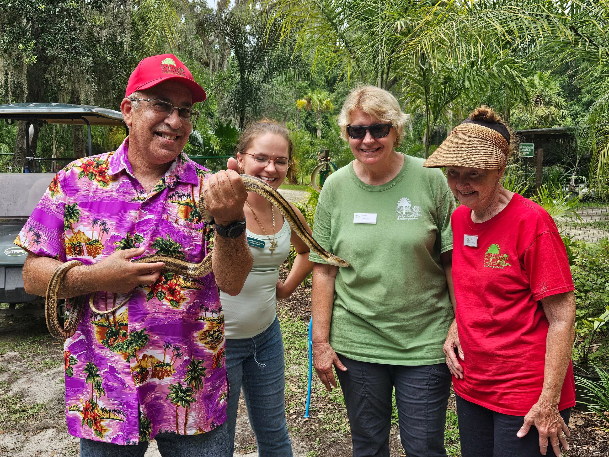 Four people outdoors, one holding a snake, smiling, surrounded by greenery, wearing casual clothing and sunglasses.