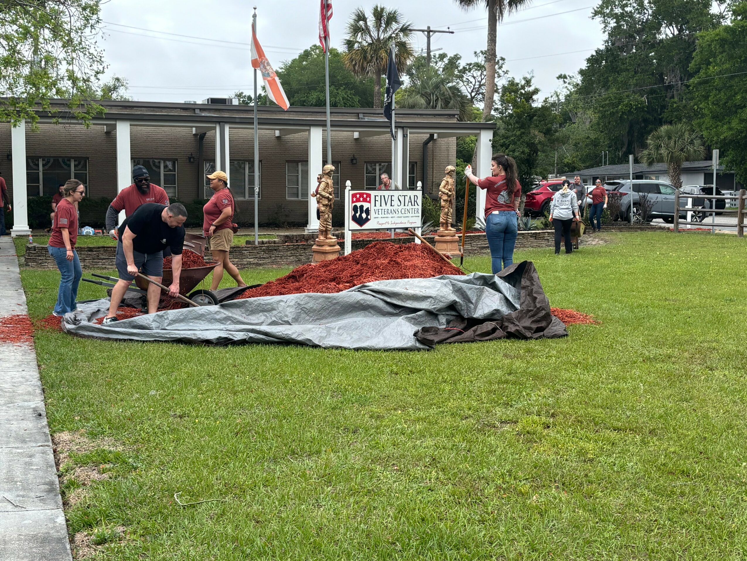 Group of people gathered around a memorial with flags, some working on a large tarp on the grass, building a structure.