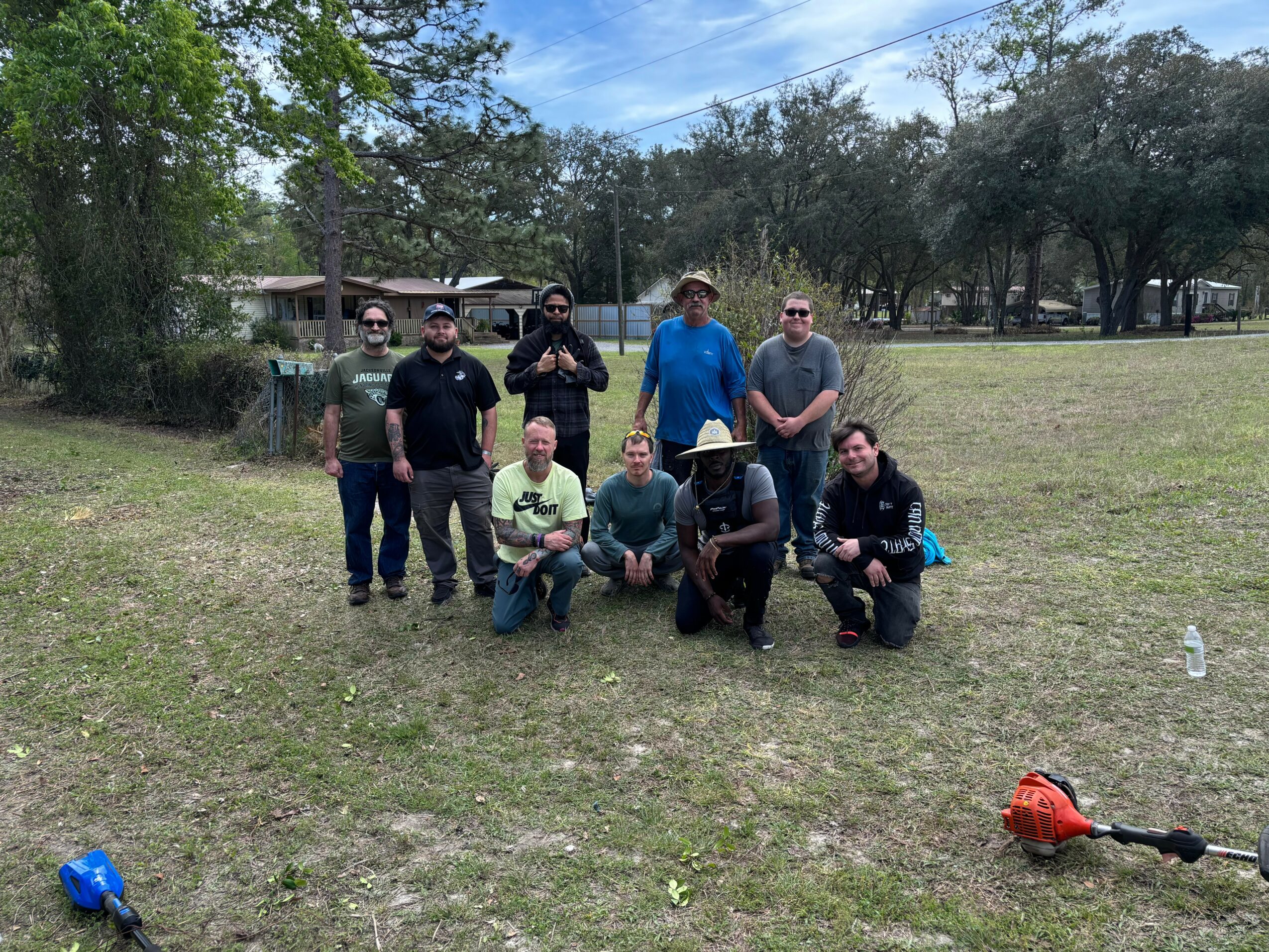 Group of nine people outdoors on grassy area, some standing and some kneeling, with trees and a house in background.