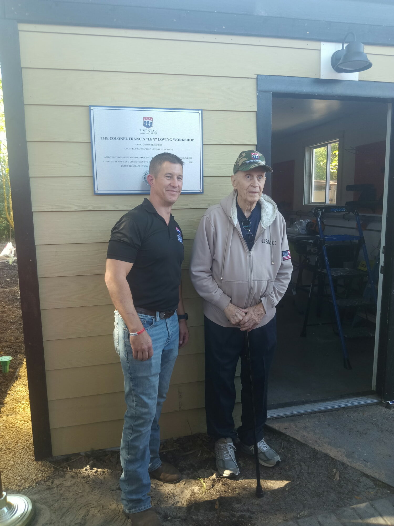 Two men standing outside a building near a door, one wearing a black shirt and jeans, the other in a jacket and cap.