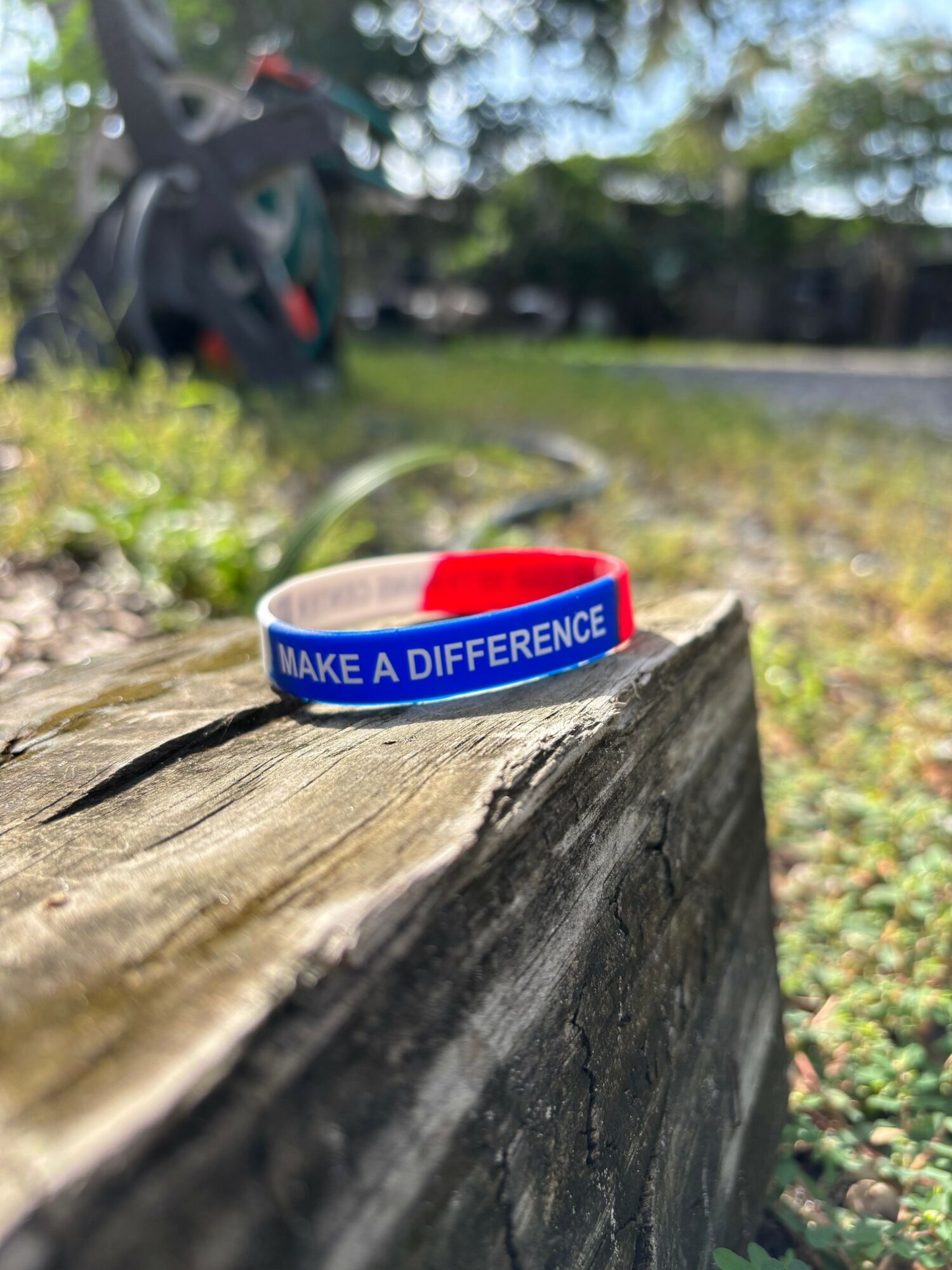 Blue and red silicone wristband with white text reading 'MAKE A DIFFERENCE' on a wooden surface outdoors.