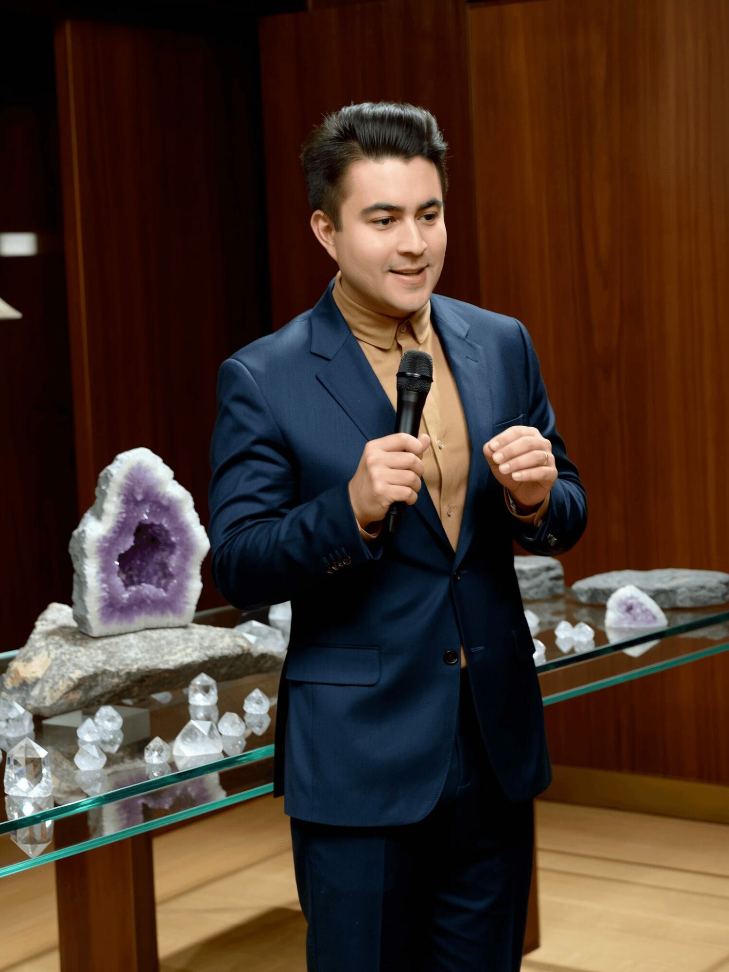 Young man in a suit holding a microphone, standing in front of a display with crystals and minerals, smiling.