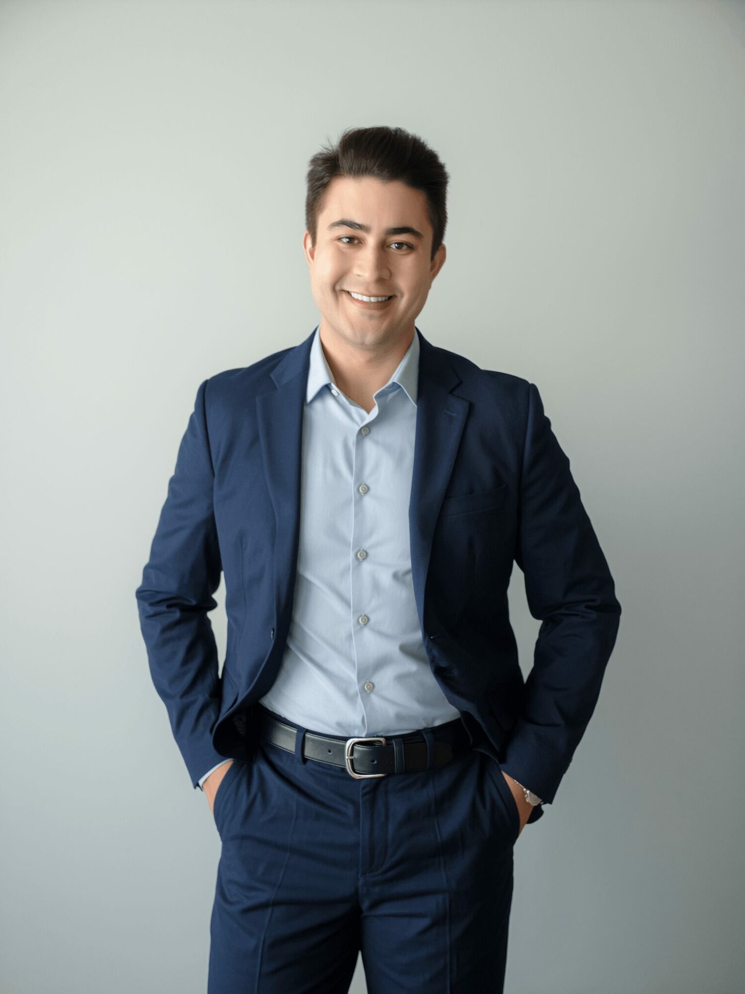 Young man in a navy suit and light blue shirt smiling with hands in pockets against a plain background.