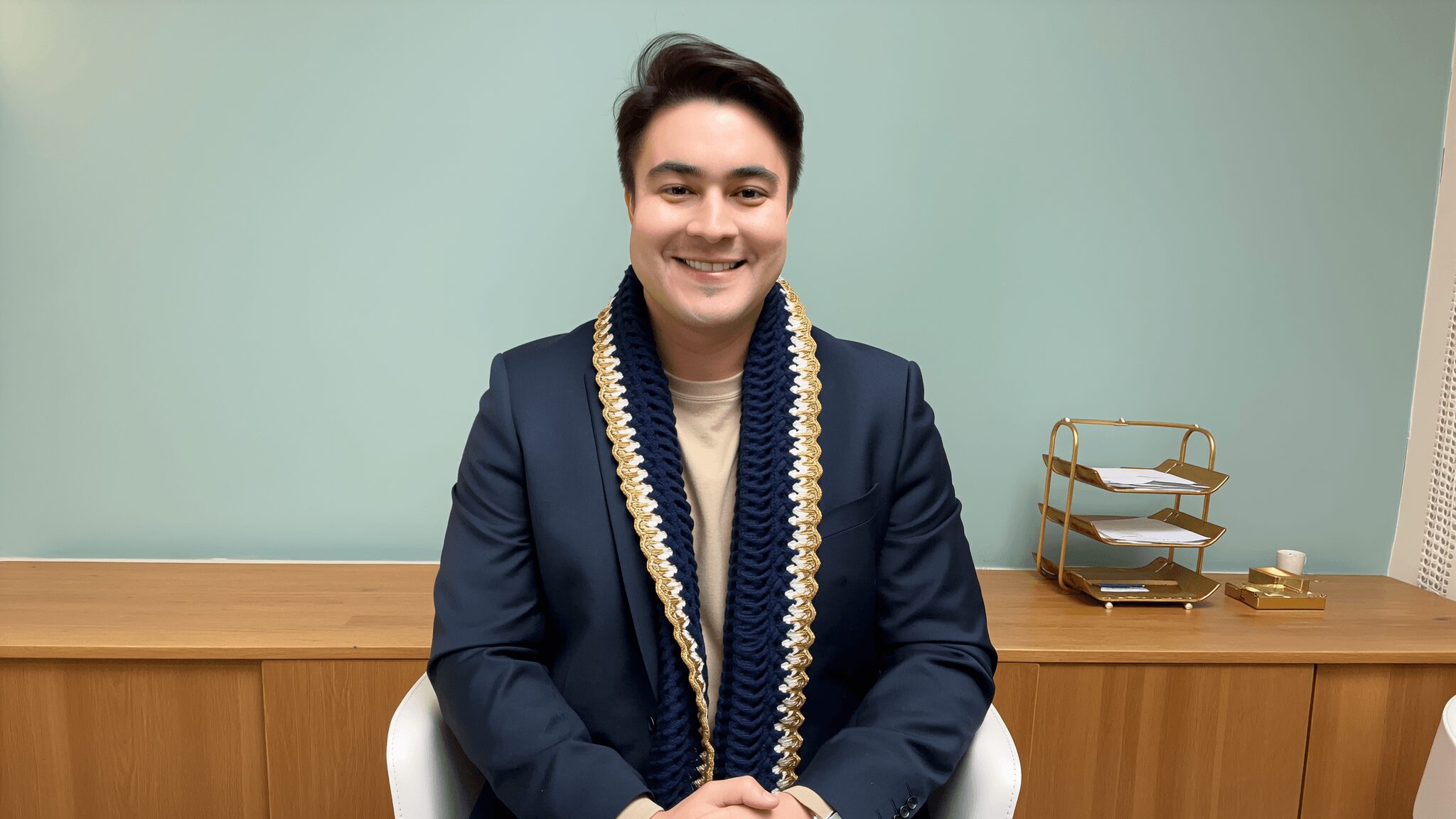 Young man sitting at a desk, smiling, wearing a dark suit and a garland, with a wooden shelf behind him.