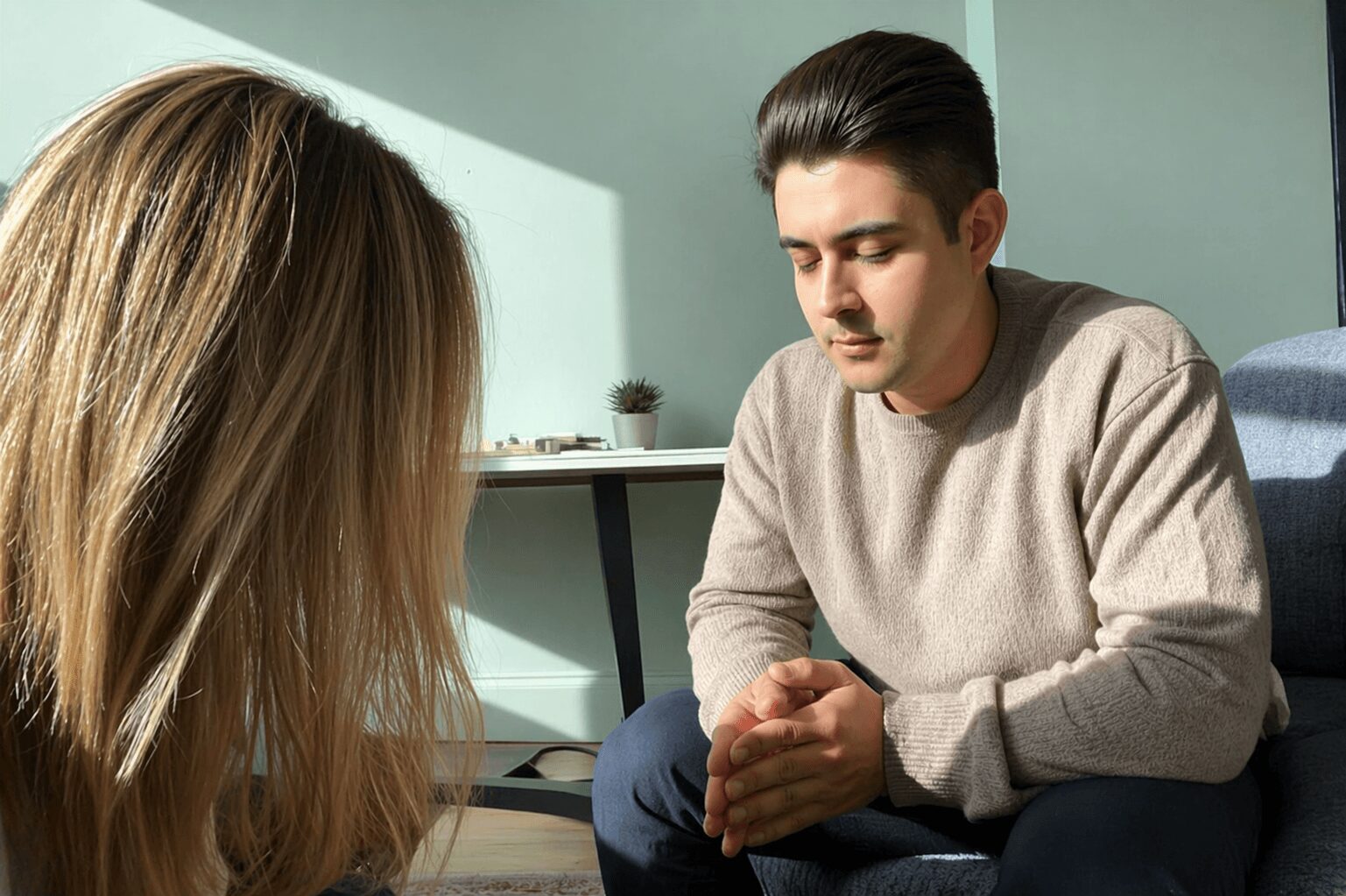 A young man and woman sit indoors, engaged in a serious conversation, with a desk and plant in the background.