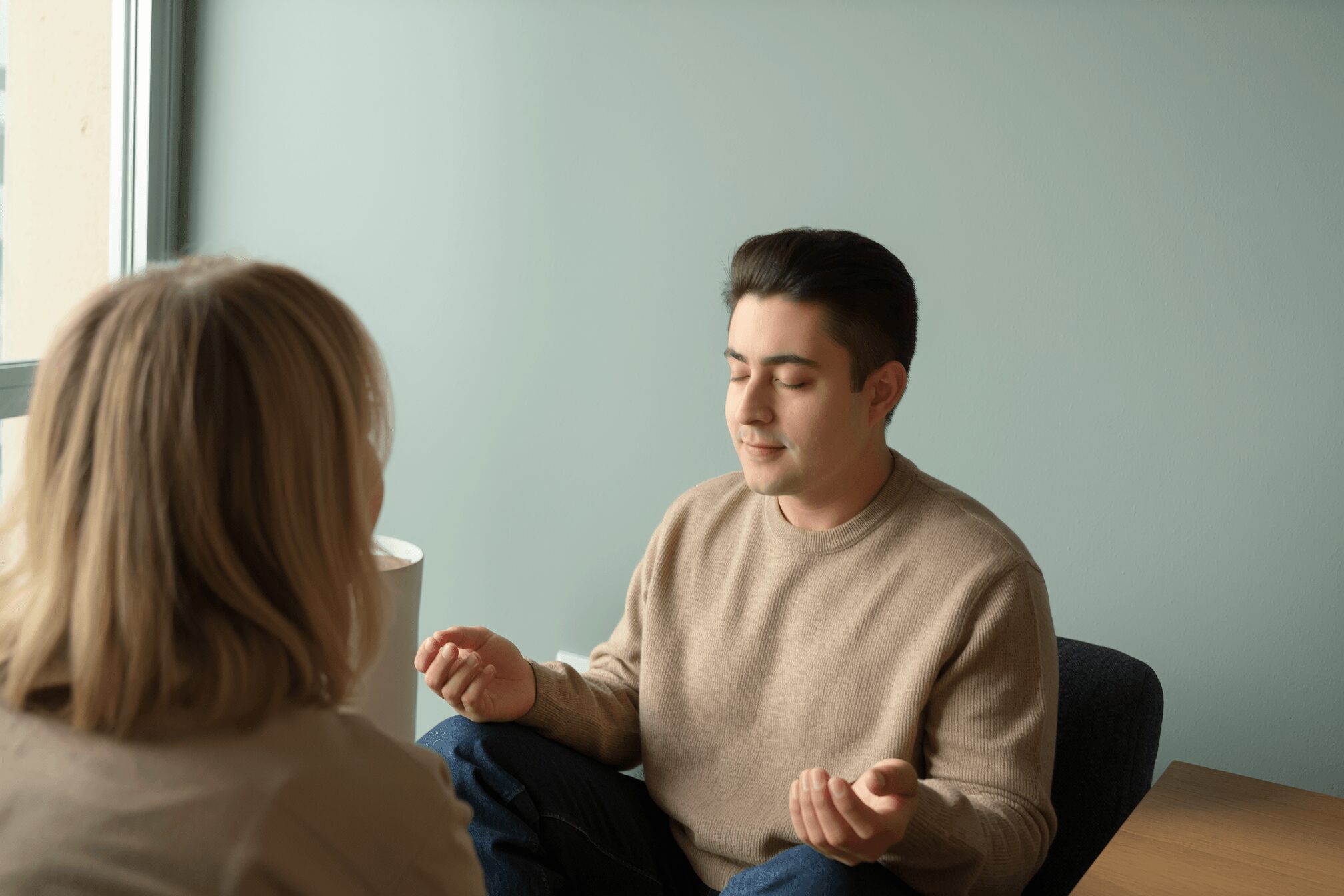 Two people sitting and talking in a room with a light green wall, one facing the camera with back to viewer.
