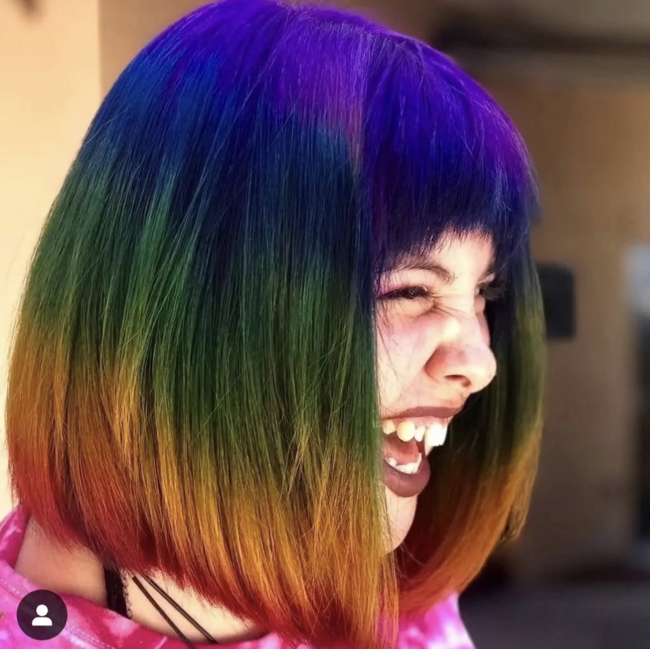 Person with a colorful rainbow hairstyle, smiling and showing teeth, with dark bangs and a pink top.