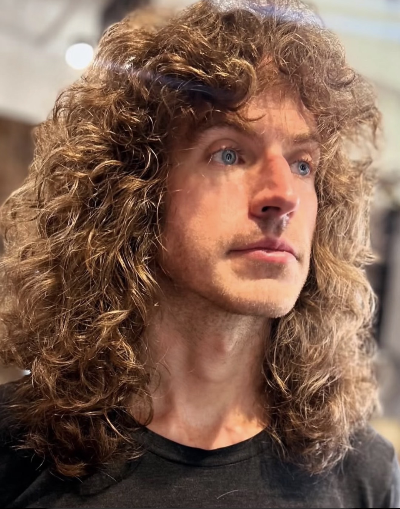 Person with long, curly hair and blue eyes looking to the side indoors.