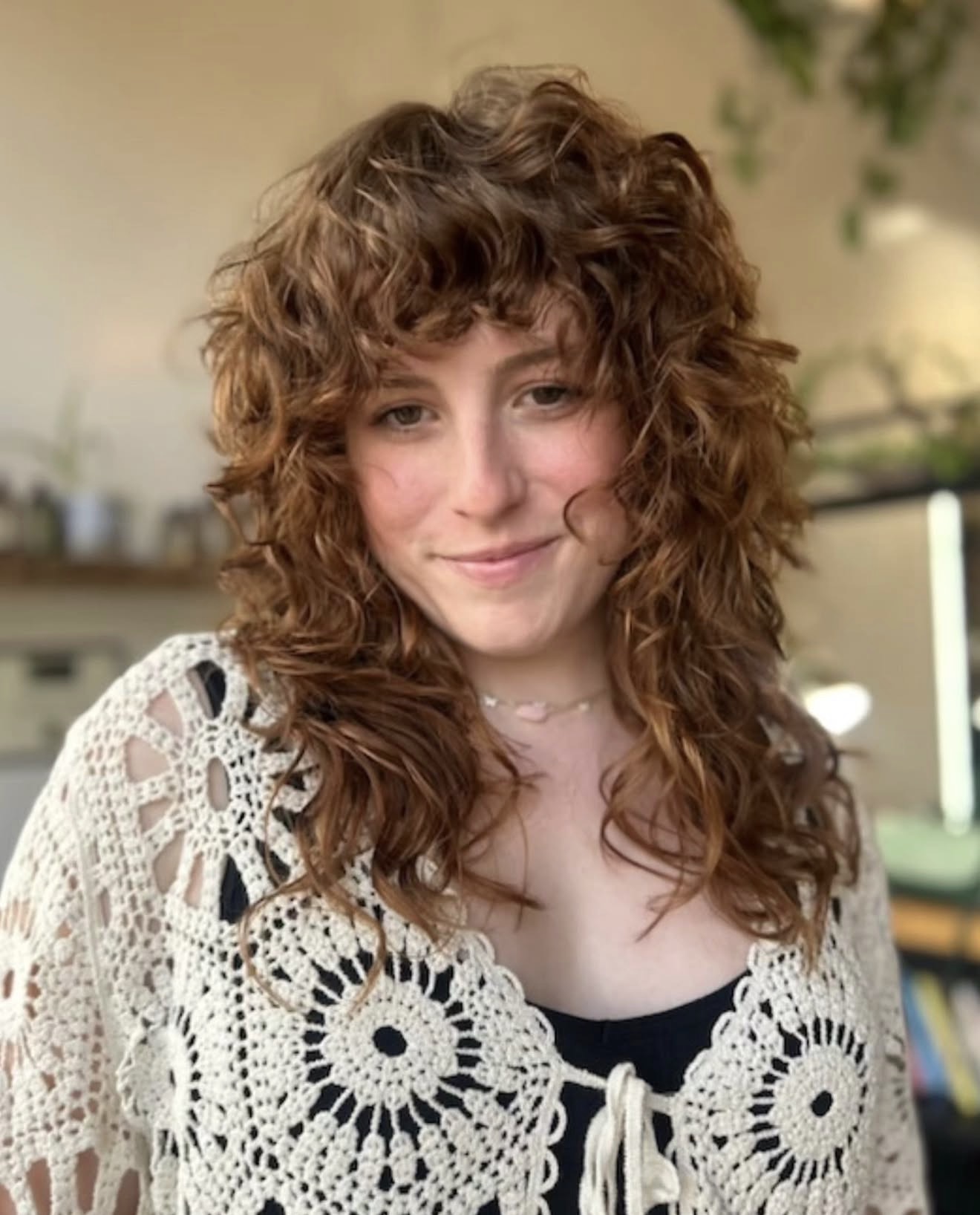 Young woman with curly hair smiling, wearing a lace top, in an indoor setting.