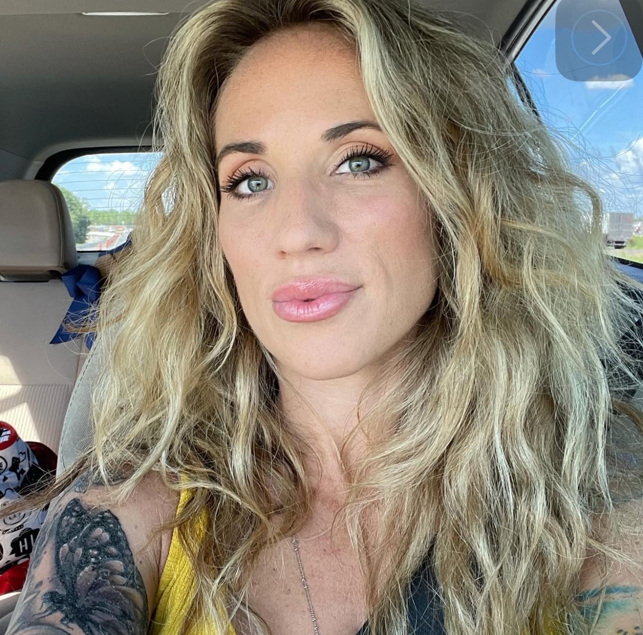 Woman with long, wavy blonde hair and blue eyes sitting in a car, wearing a yellow top.