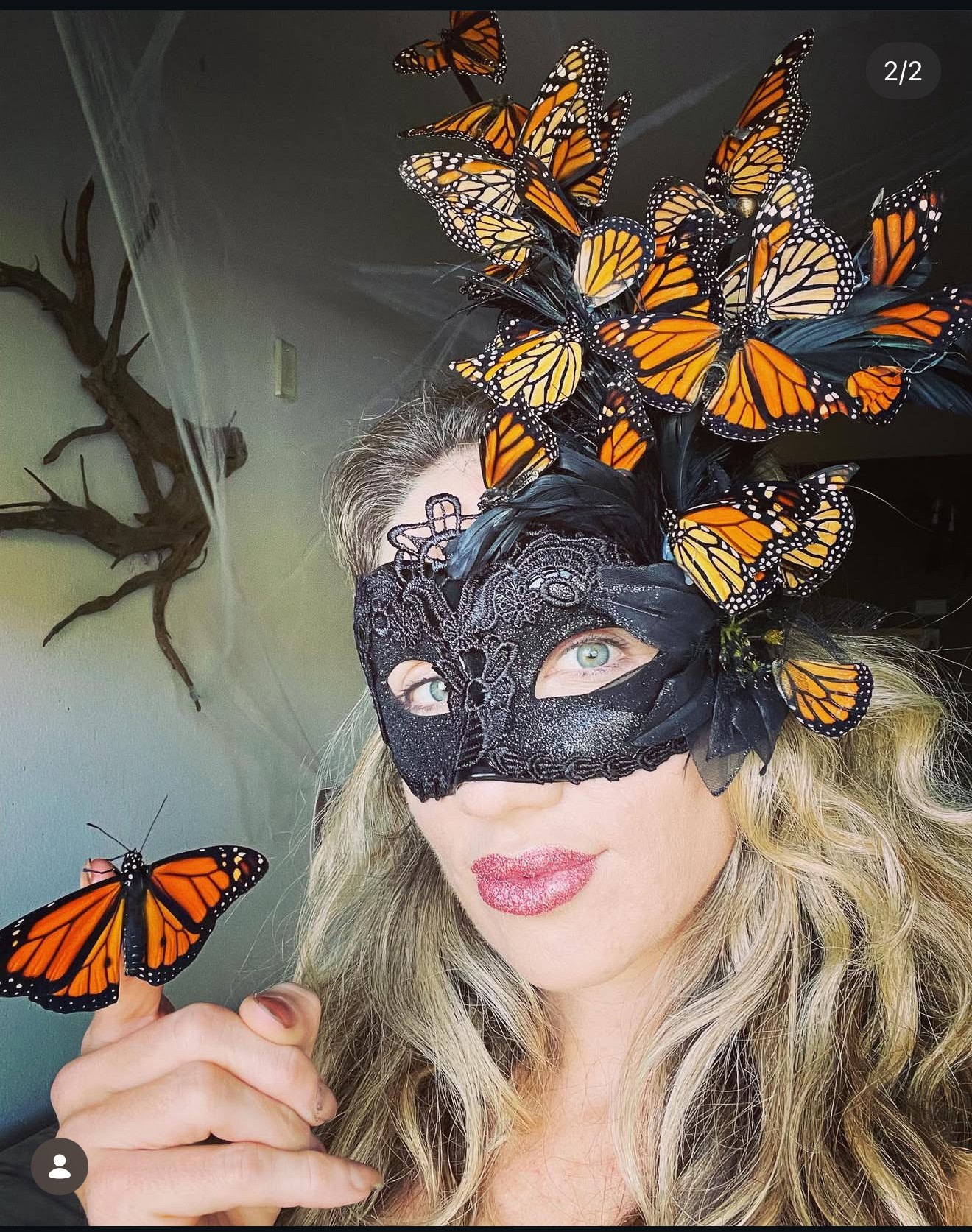 Woman wearing a black lace mask with numerous orange and black butterflies on her head and one butterfly on her finger.