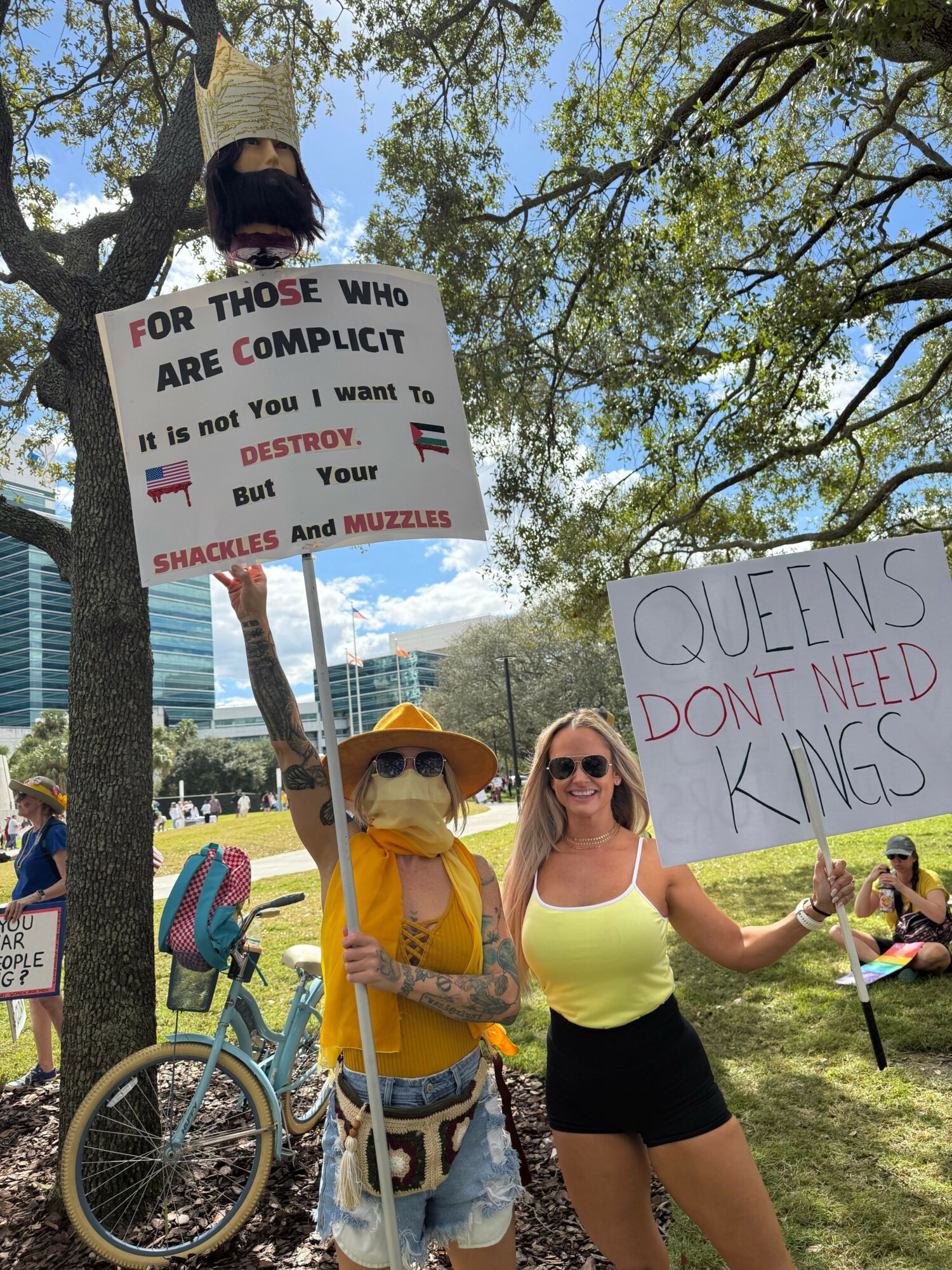Two women holding protest signs outdoors, one on a bicycle, trees and a park in the background.