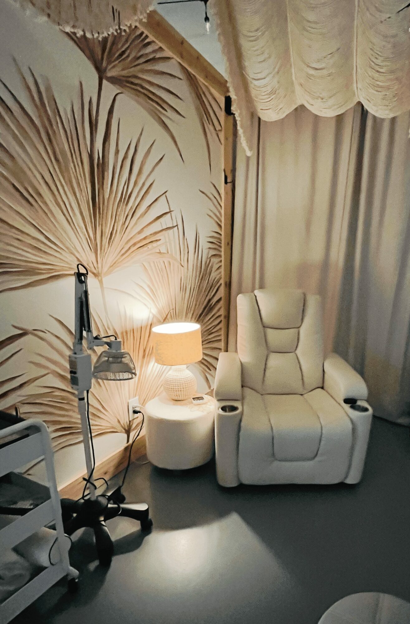 Comfortable white recliner chair next to a small table with a lamp, in a room with curtains and a decorative wall.