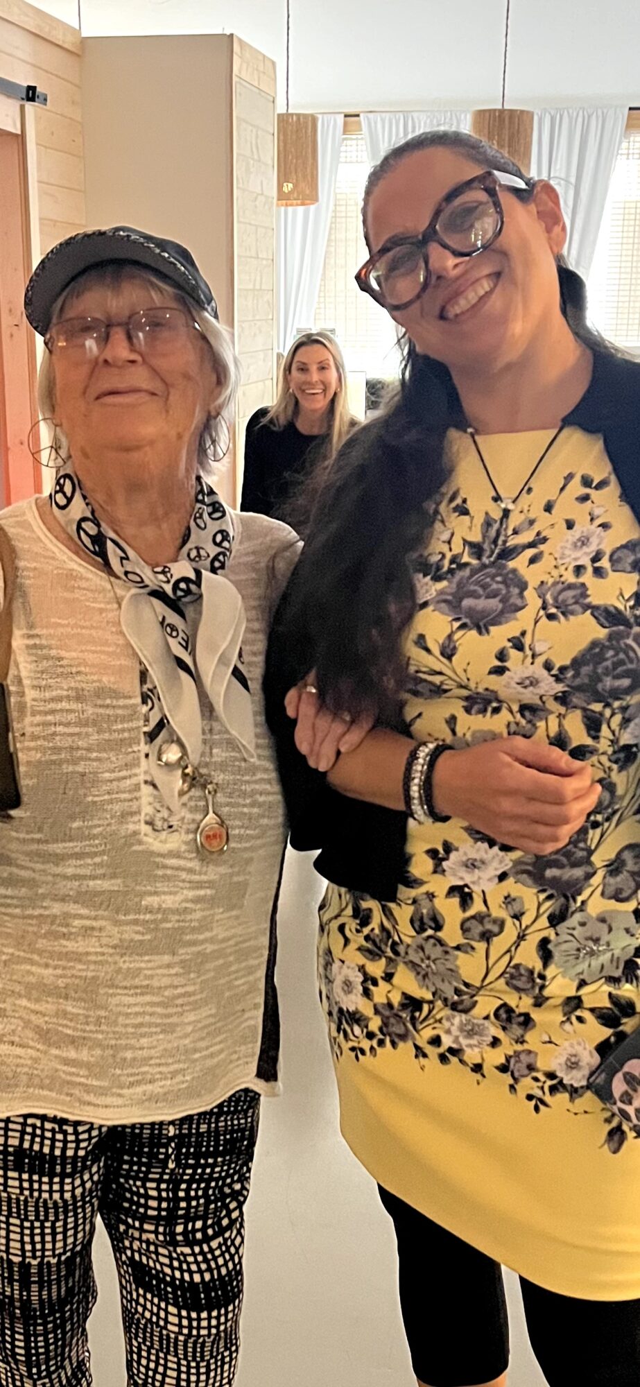 Two women standing indoors, smiling, with a woman in the background. One woman wears glasses, a hat, and patterned pants.
