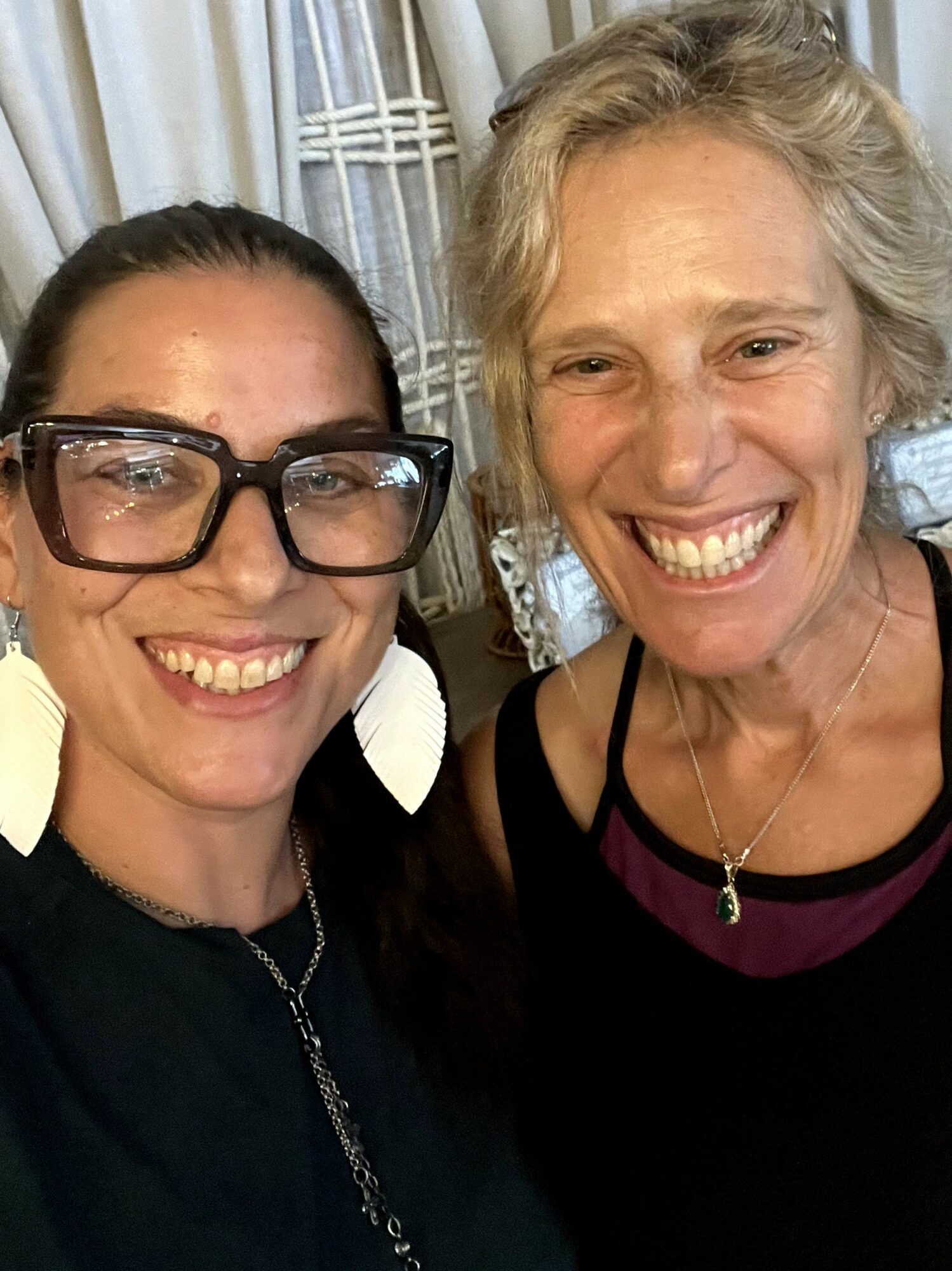 Two women smiling, one with glasses and earrings, the other with short gray hair, both in dark clothing.