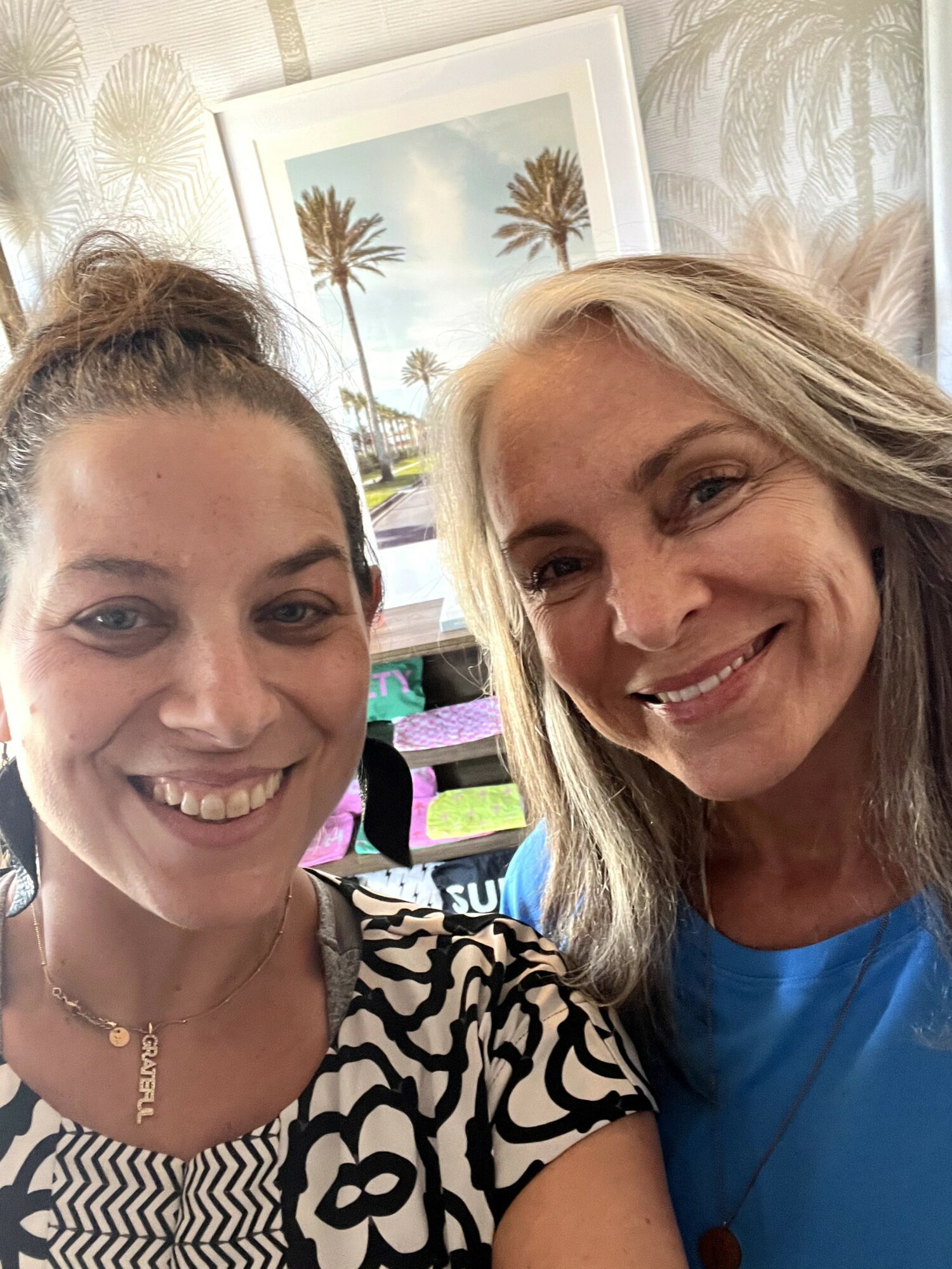 Two women smiling, taking a selfie indoors with a framed picture of palm trees in the background.