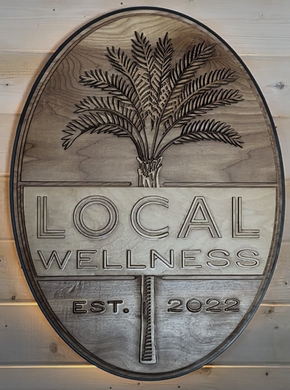 Oval sign with a palm tree illustration and text: 'LOCAL WELLNESS EST. 2022' on a wooden background.