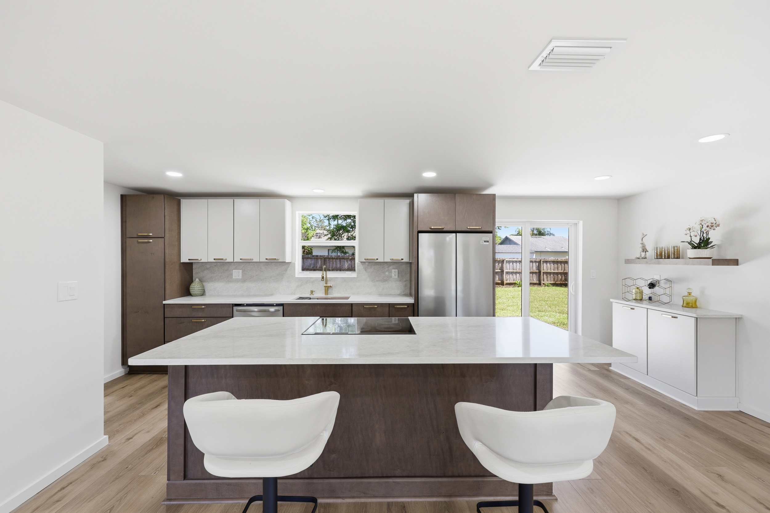 Modern kitchen with white and brown cabinets, island with two chairs, stainless steel refrigerator, and a door leading outside.