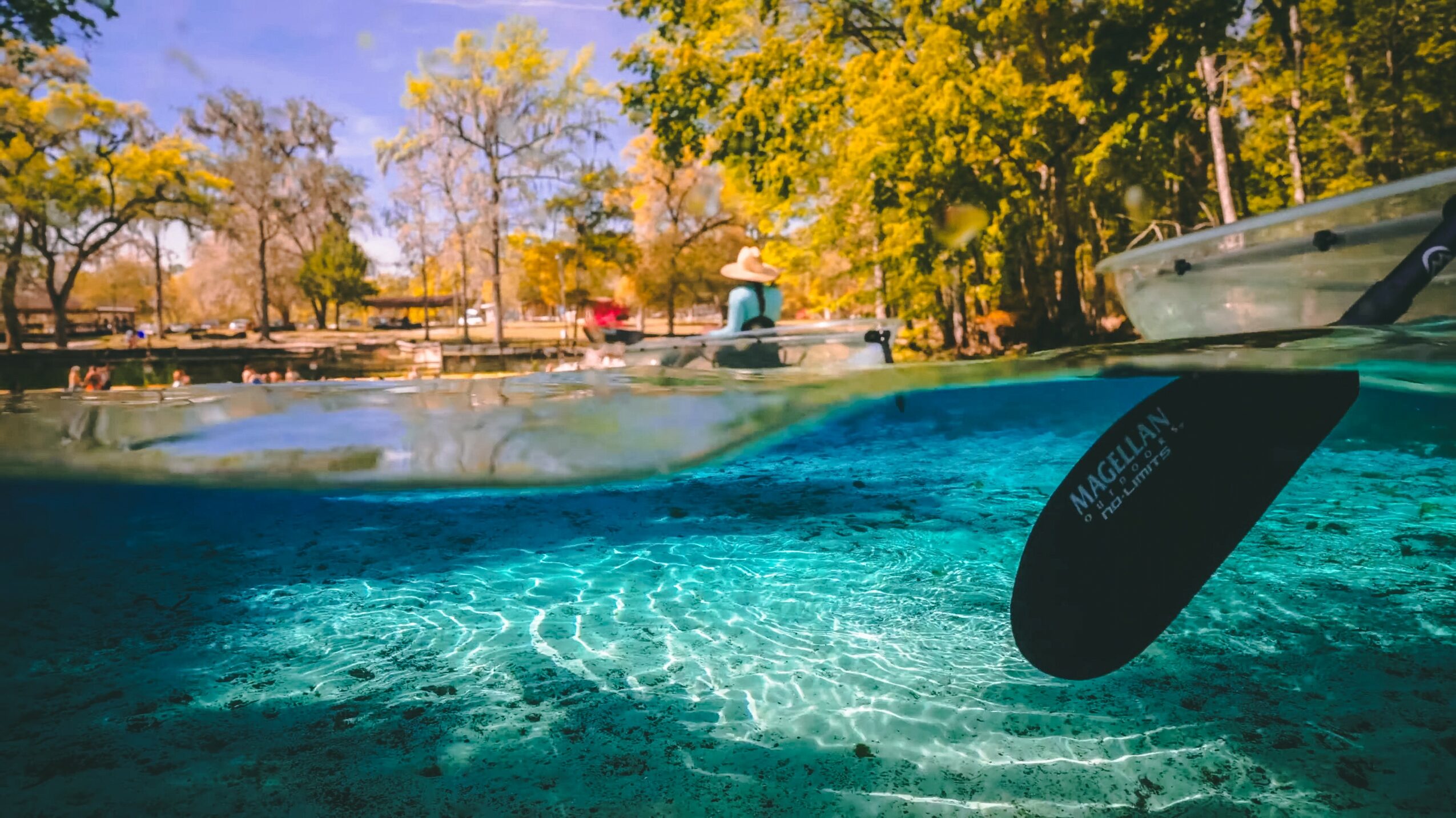 Underwater view of a kayak paddle and water, with trees and a person on the shore in the background.