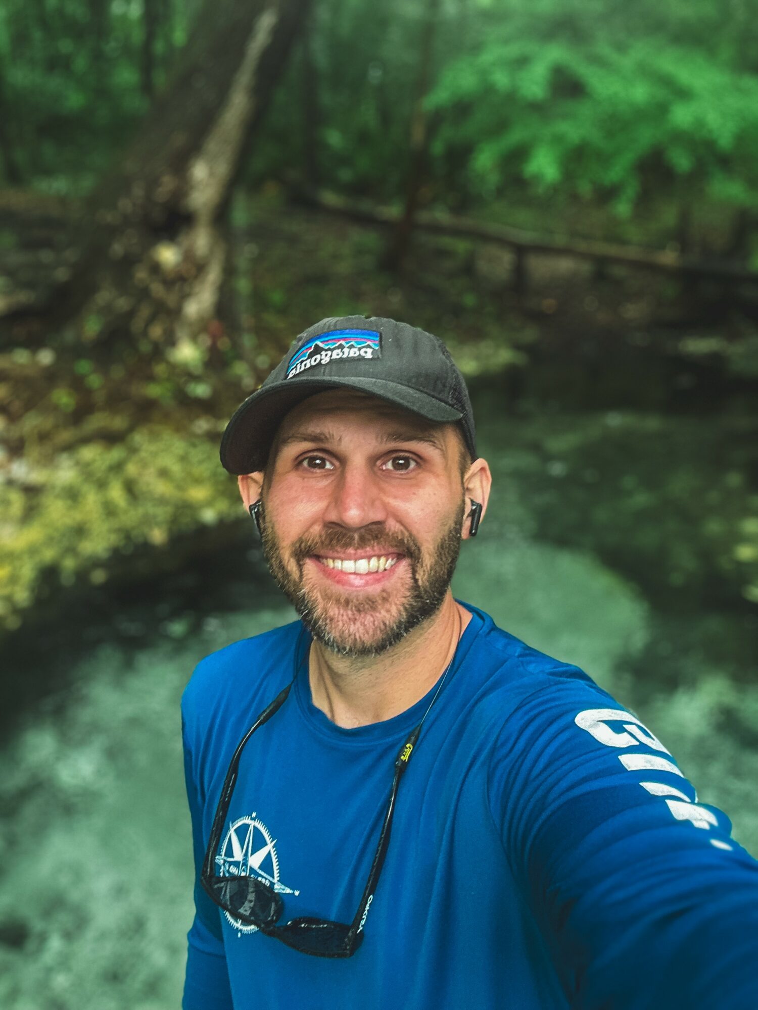 Man with a beard and cap smiling outdoors in a forested area.