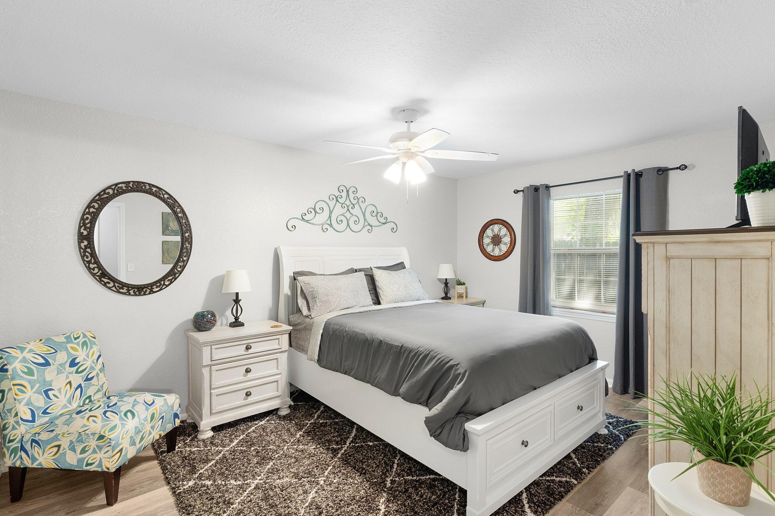 Bedroom with bed, nightstands, armchair, window, and decorative wall mirror, with ceiling fan and potted plant.