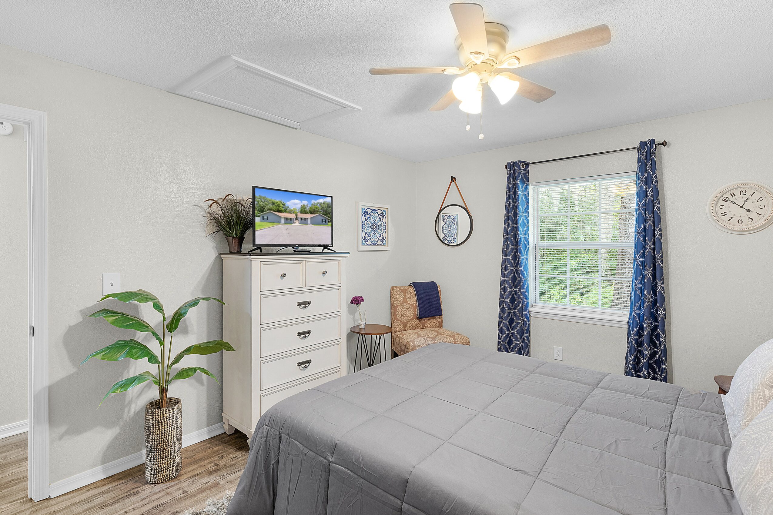 Bedroom with bed, dresser, window with blue curtains, ceiling fan, and wall clock.