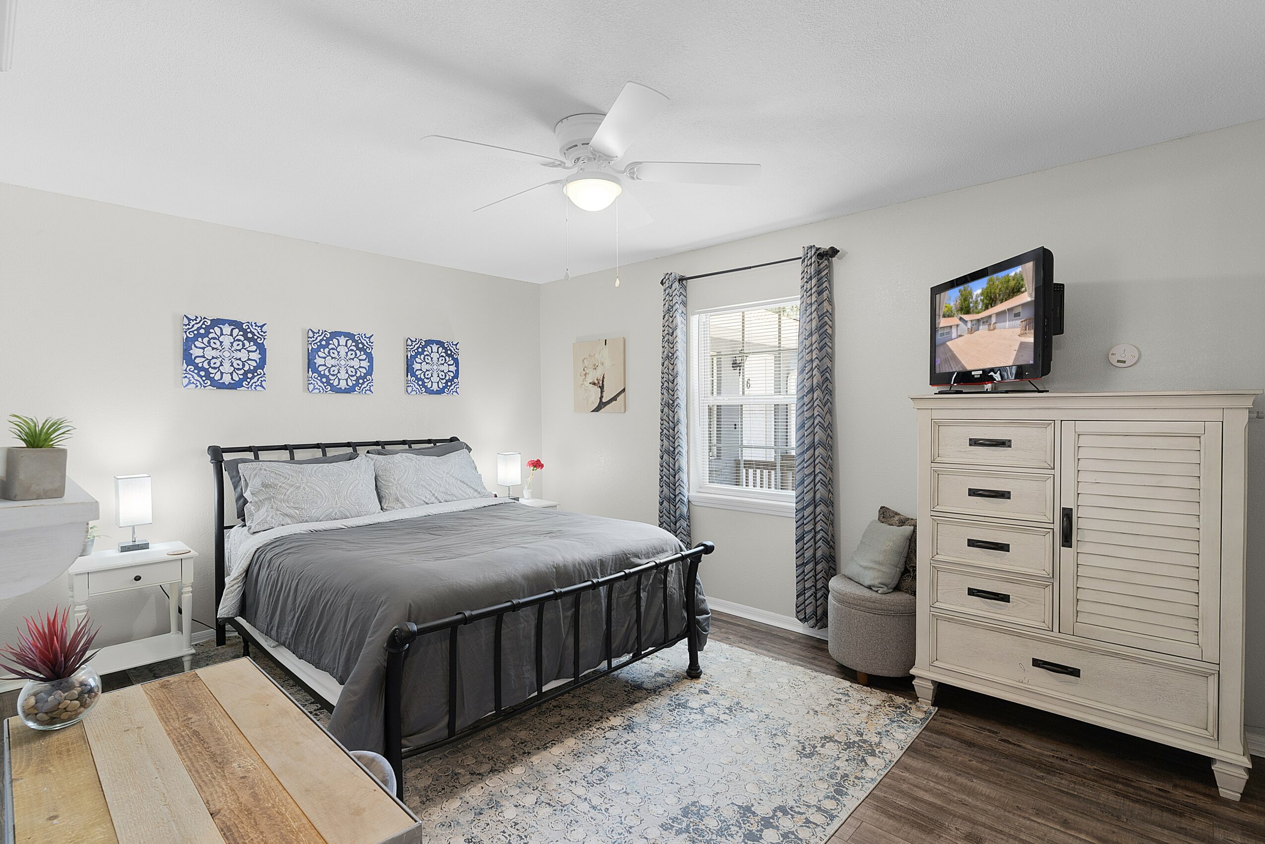 Bedroom with bed, dresser, TV, window, and ceiling fan, decorated with curtains, artwork, and a rug.