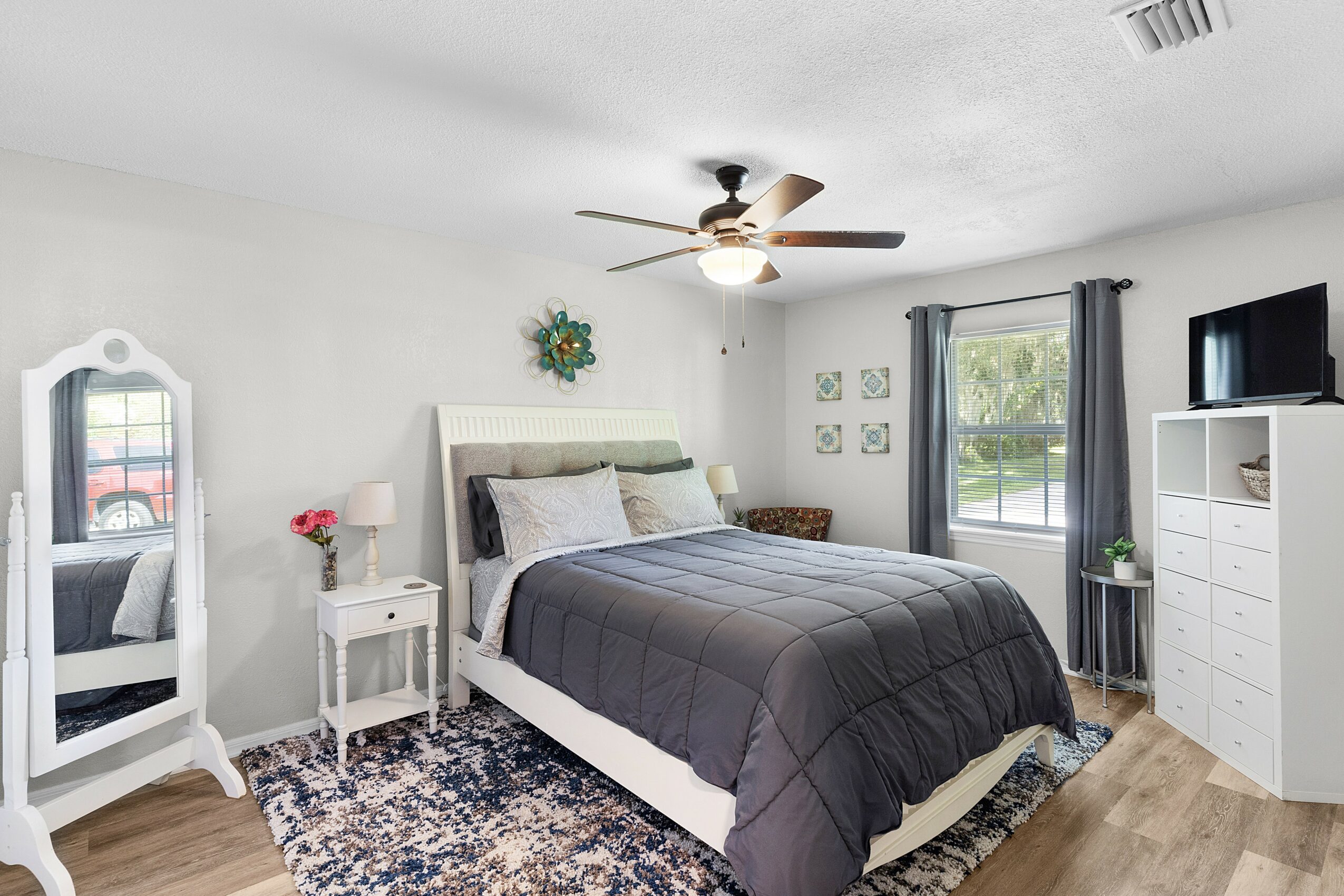 Bedroom with bed, nightstand, mirror, dresser, window with curtains, ceiling fan, and hardwood floor.