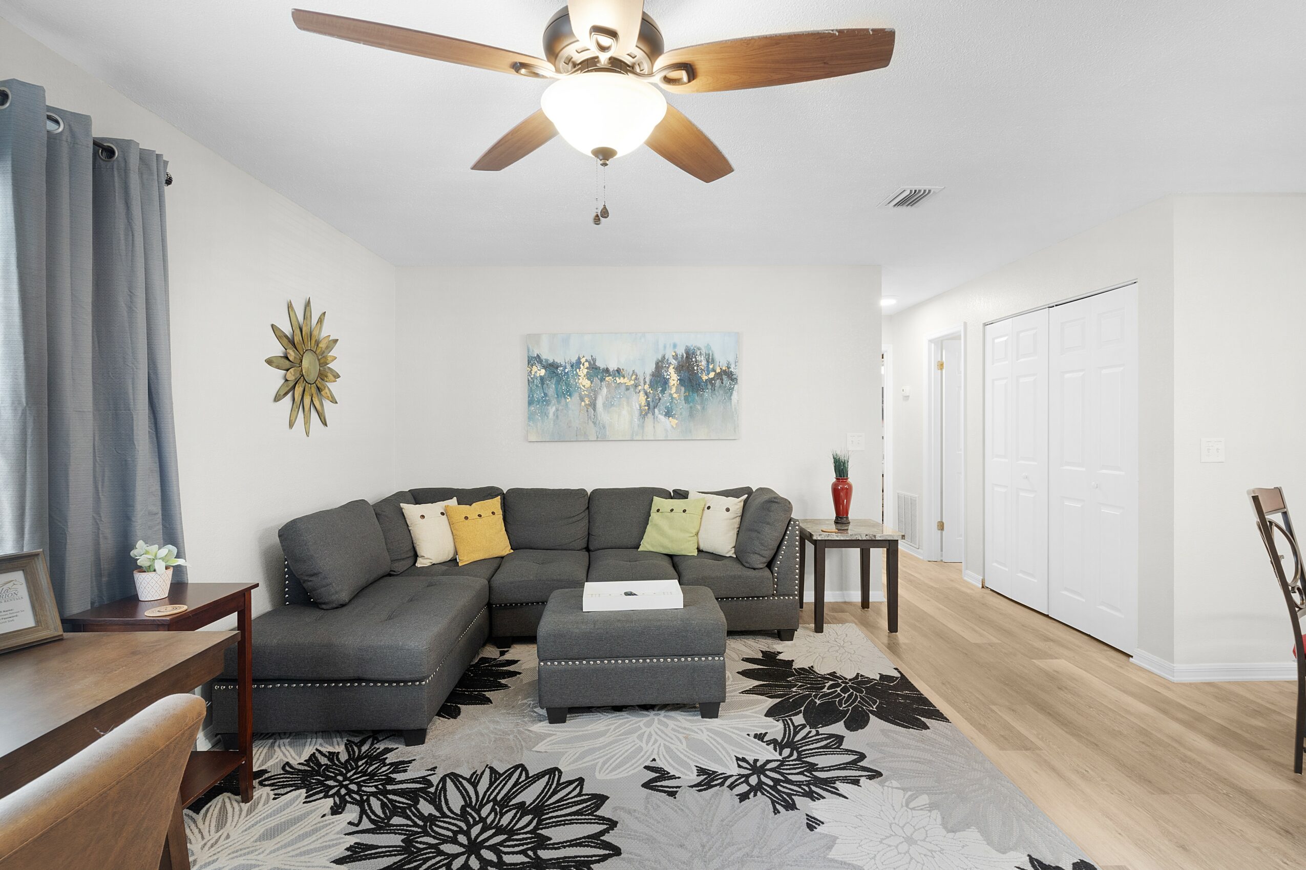 Living room with gray sectional sofa, yellow and green pillows, artwork, ceiling fan, and wooden furniture.