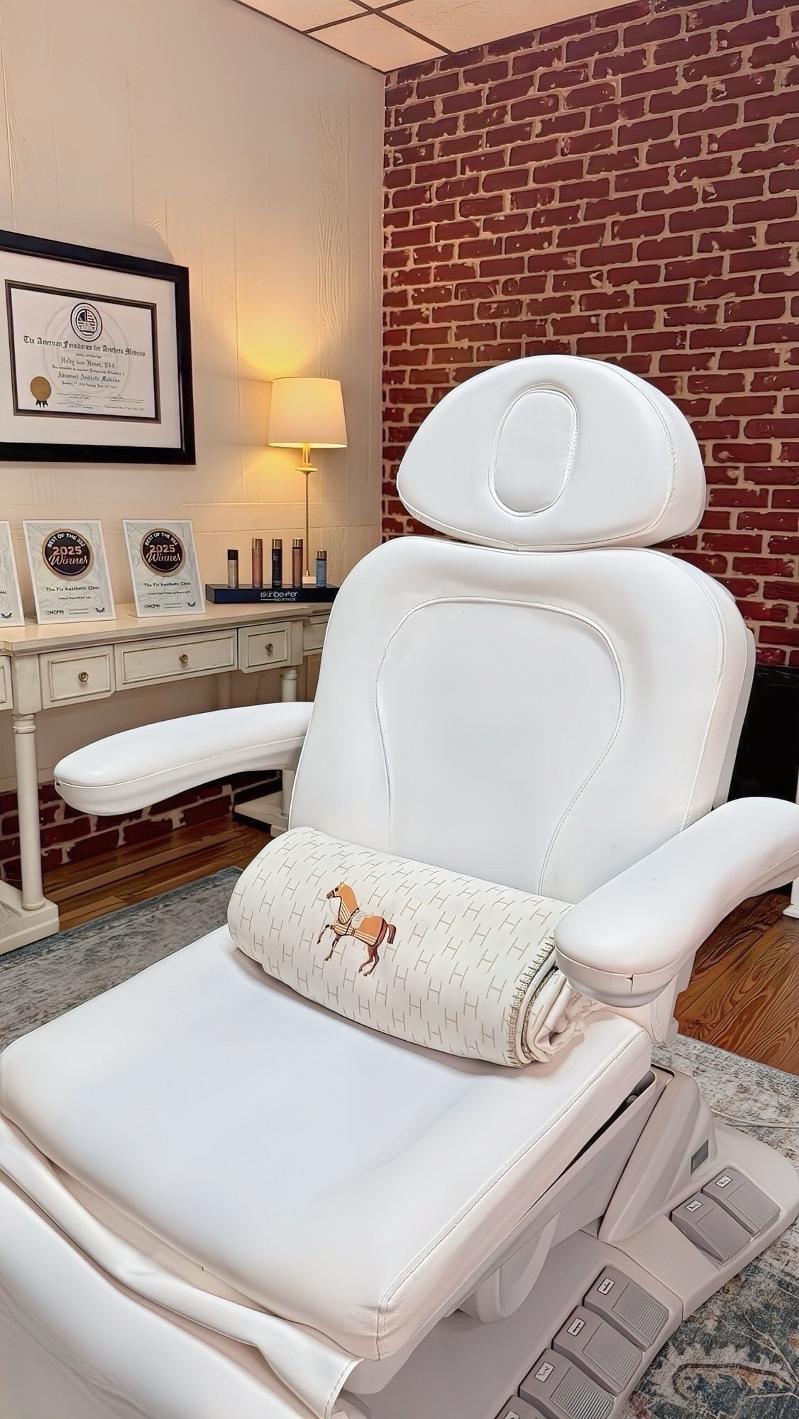 Medical or massage chair with a pillow featuring a horse illustration, in a room with brick wall and framed certificates.