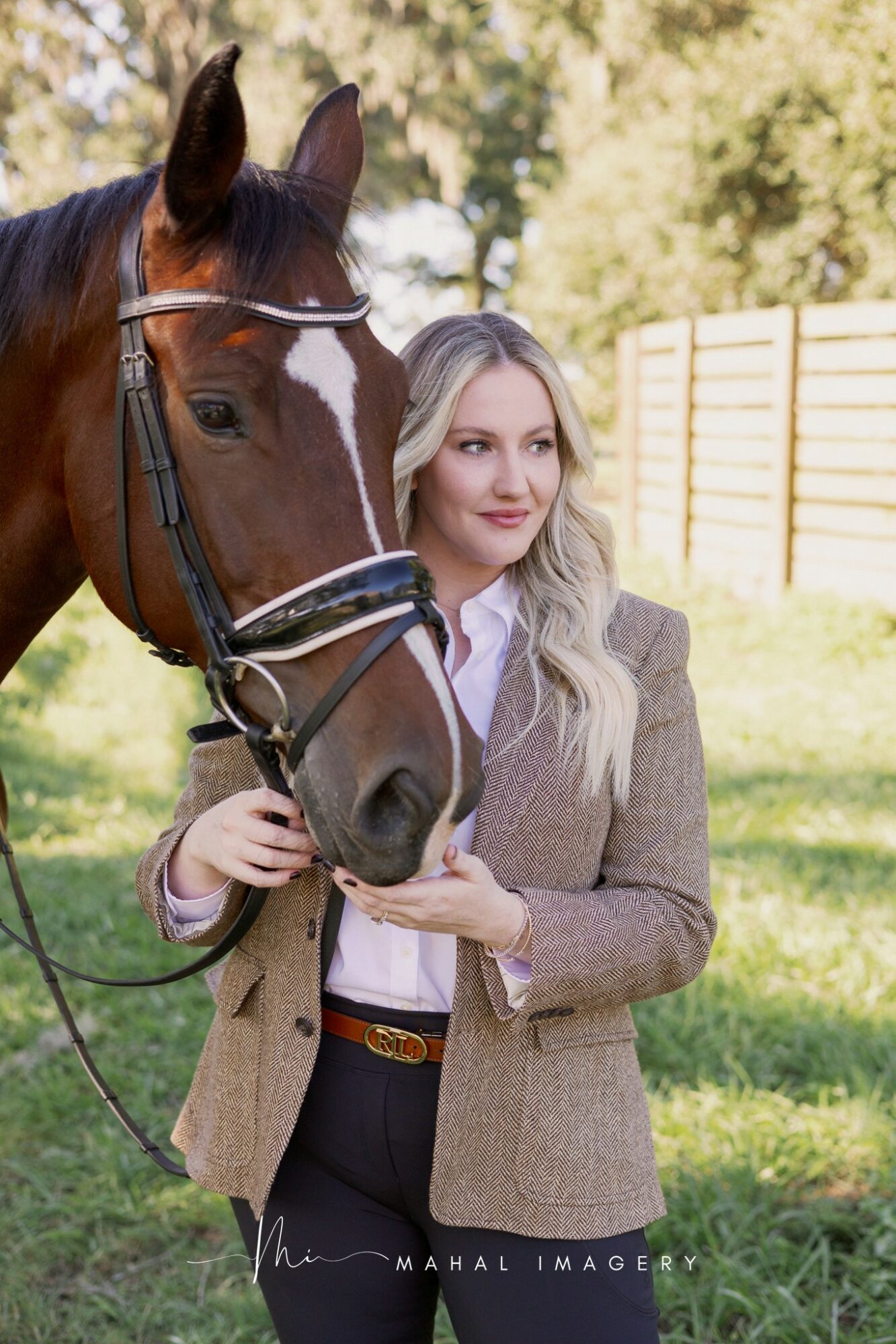 Woman with long blonde hair standing beside a brown horse outdoors, holding its bridle, with trees and a fence in background.