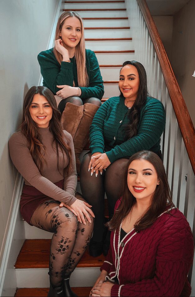 Four women sitting on staircase, smiling, indoors, casual clothing, wooden staircase, railing, background wall.