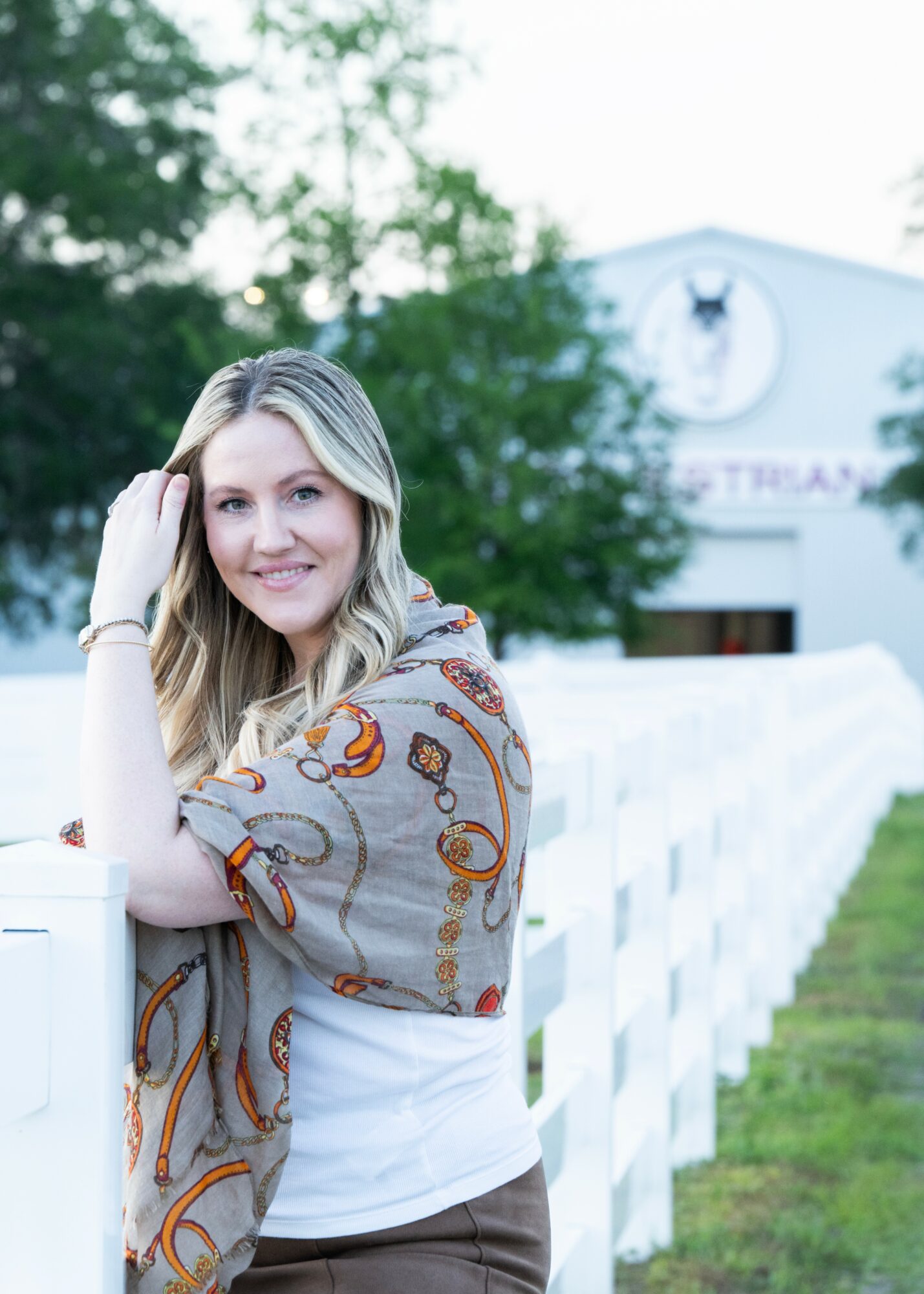 Young woman with blonde hair smiling, leaning on a white fence outdoors, with trees and a building in background.