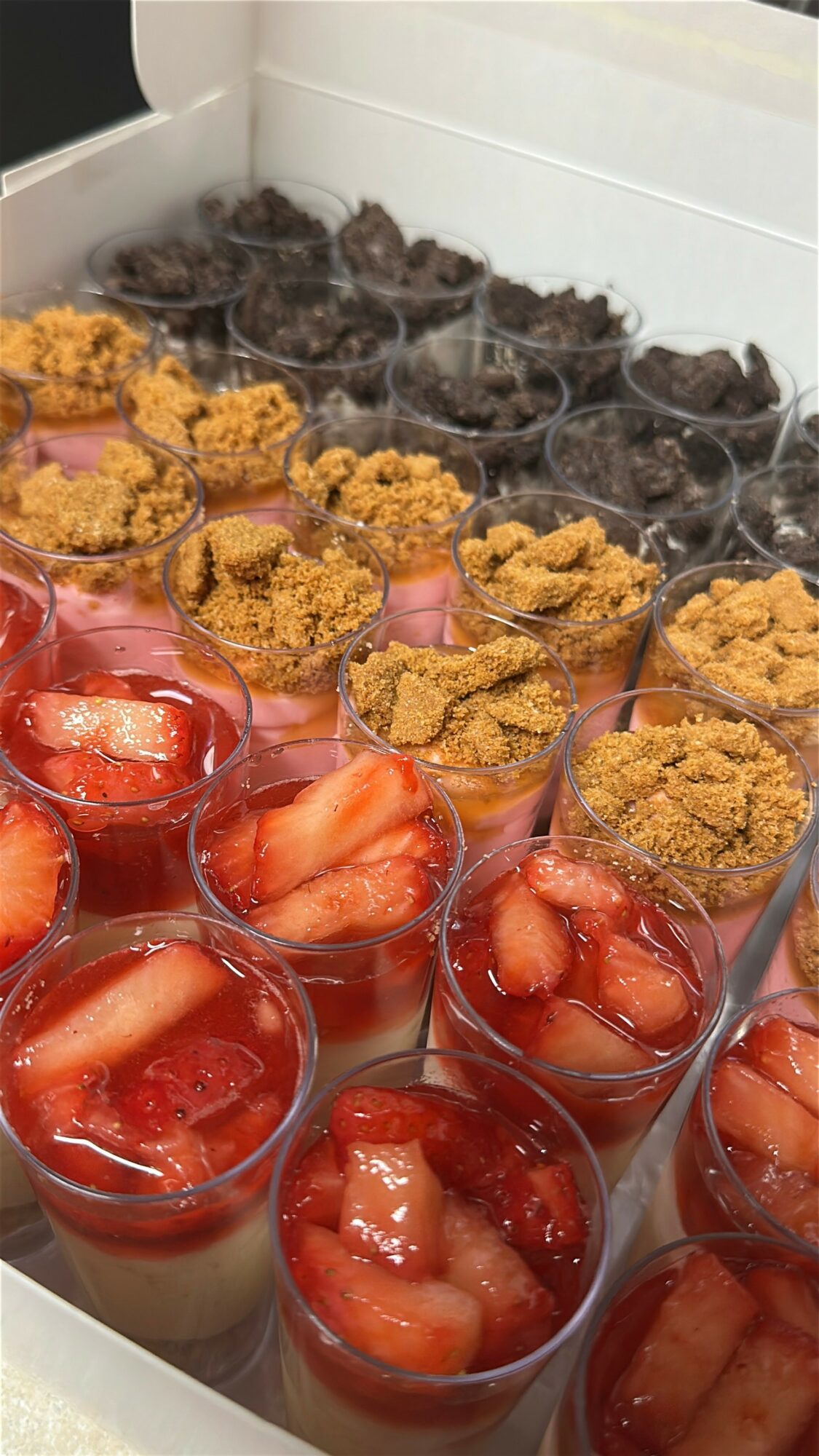 Assorted desserts in cups with strawberries, crumb topping, and chocolate pieces in a box.