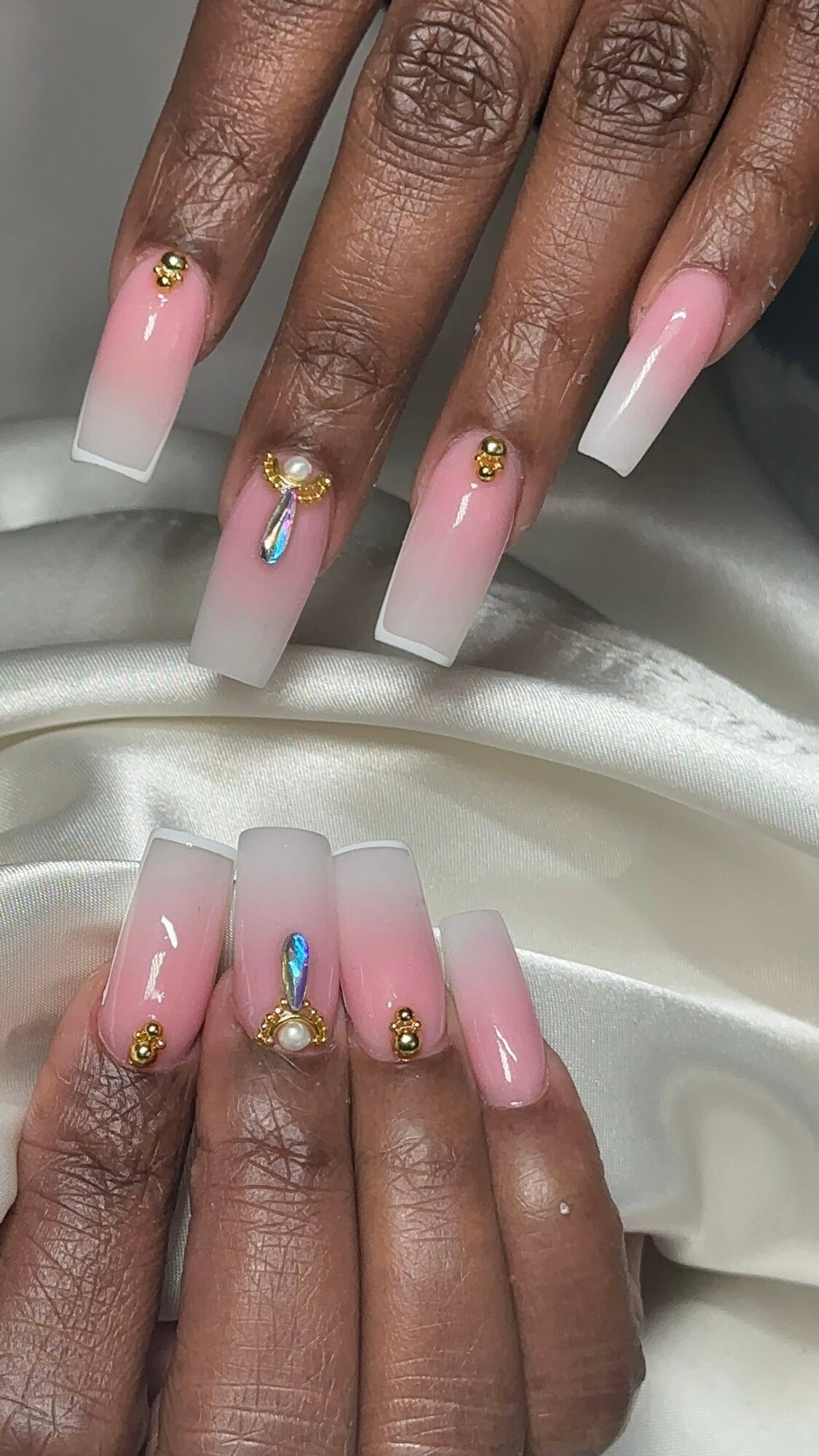 Hands with long, square-shaped nails painted in pink and white gradient, decorated with gold and blue embellishments.