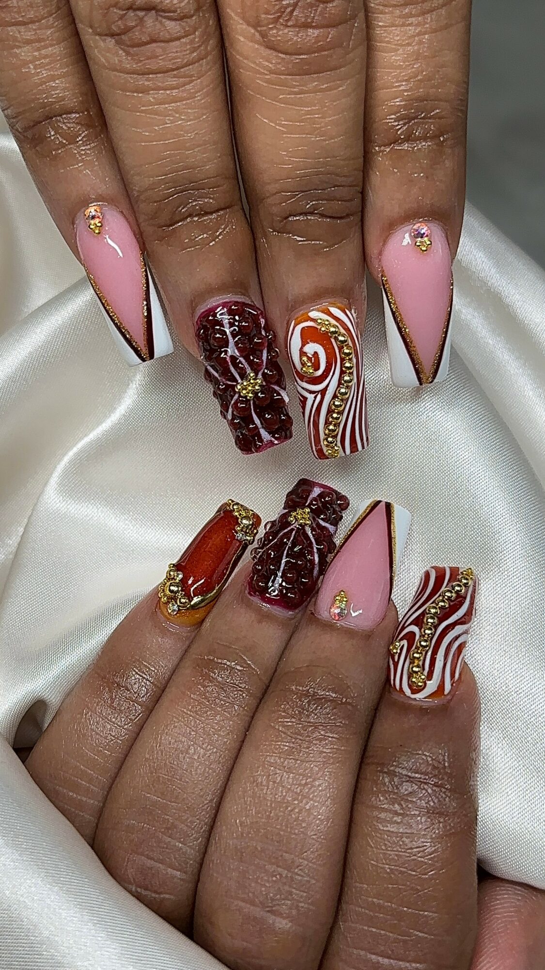 Nail art with pink, red, gold, and white patterns, including swirls, marbling, and embellishments on long nails.