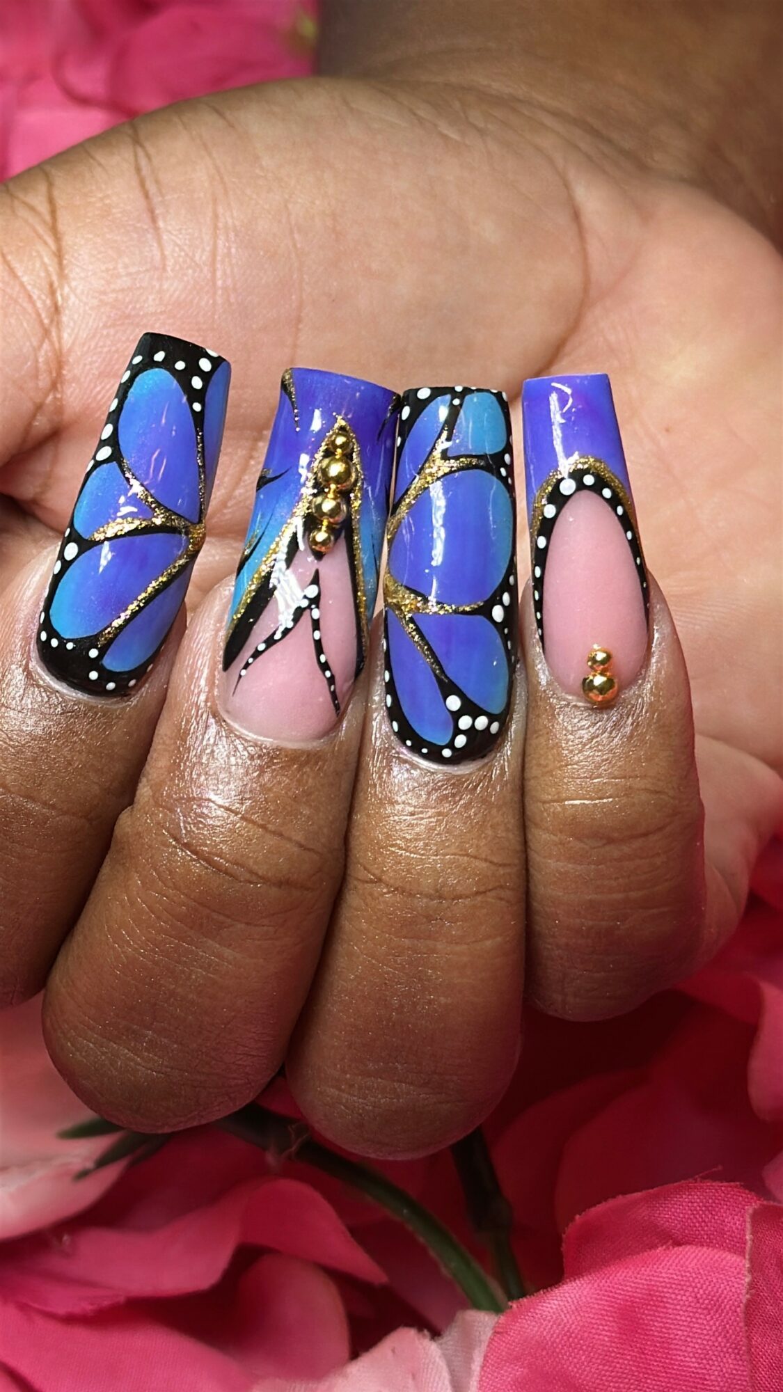 Close-up of decorated fingernails with butterfly and floral designs, pink and blue colors, gold accents, and rhinestones.