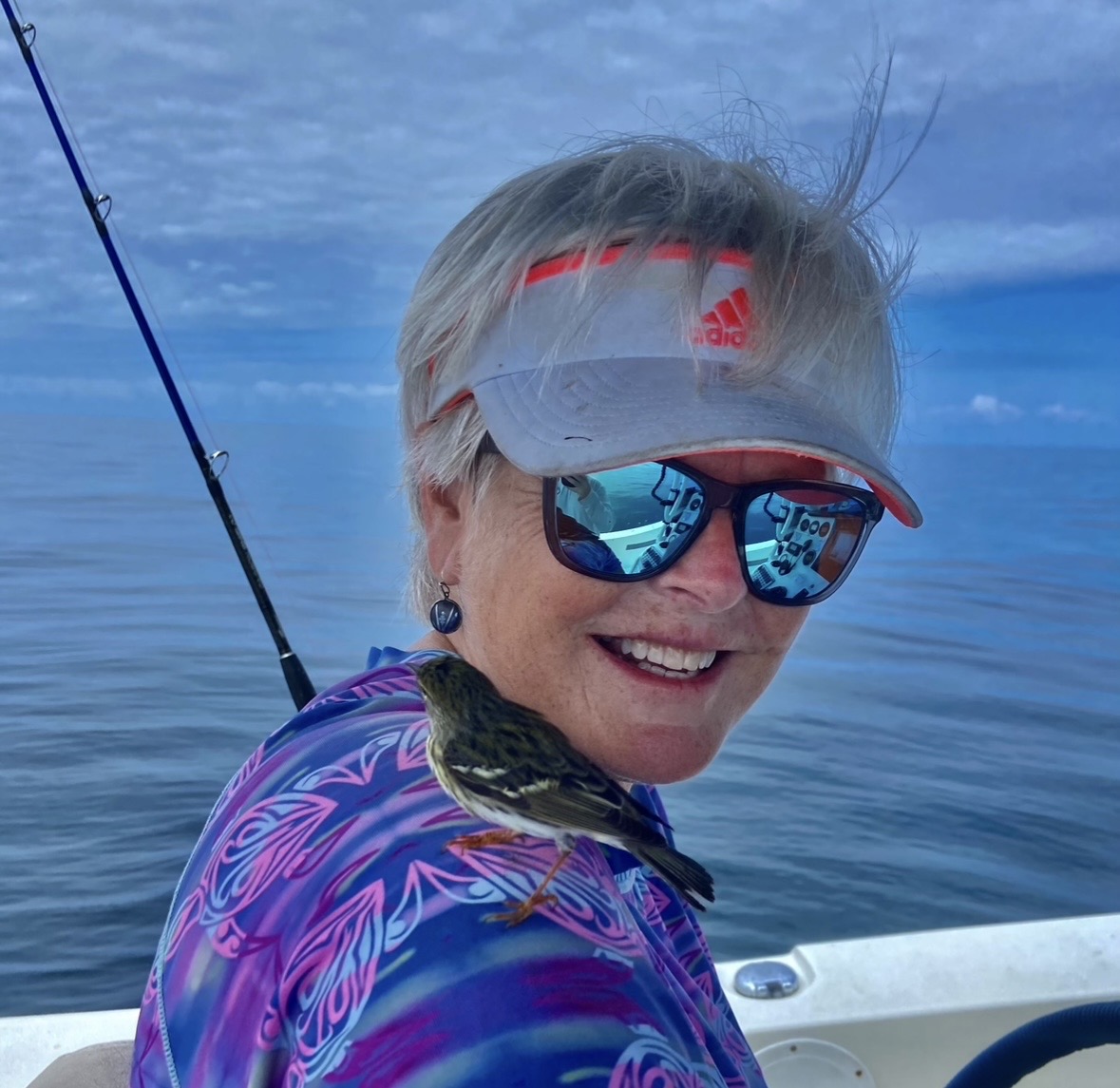 Smiling woman wearing sunglasses, a visor, and a colorful jacket on a boat with water background.