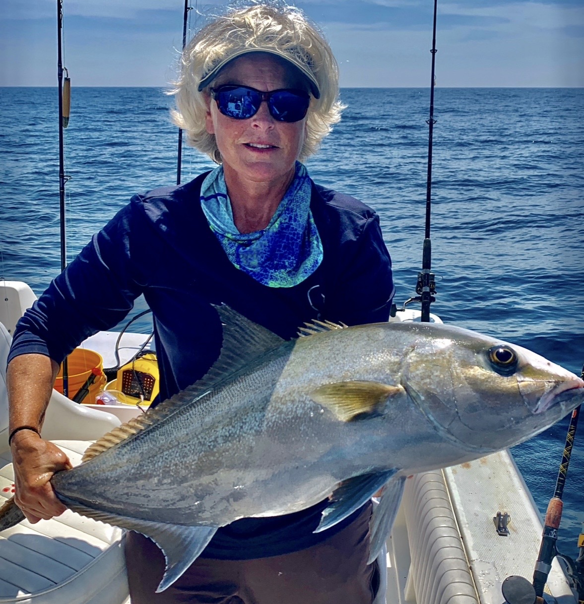 Woman holding large fish on boat with ocean background, wearing sunglasses and a blue scarf.