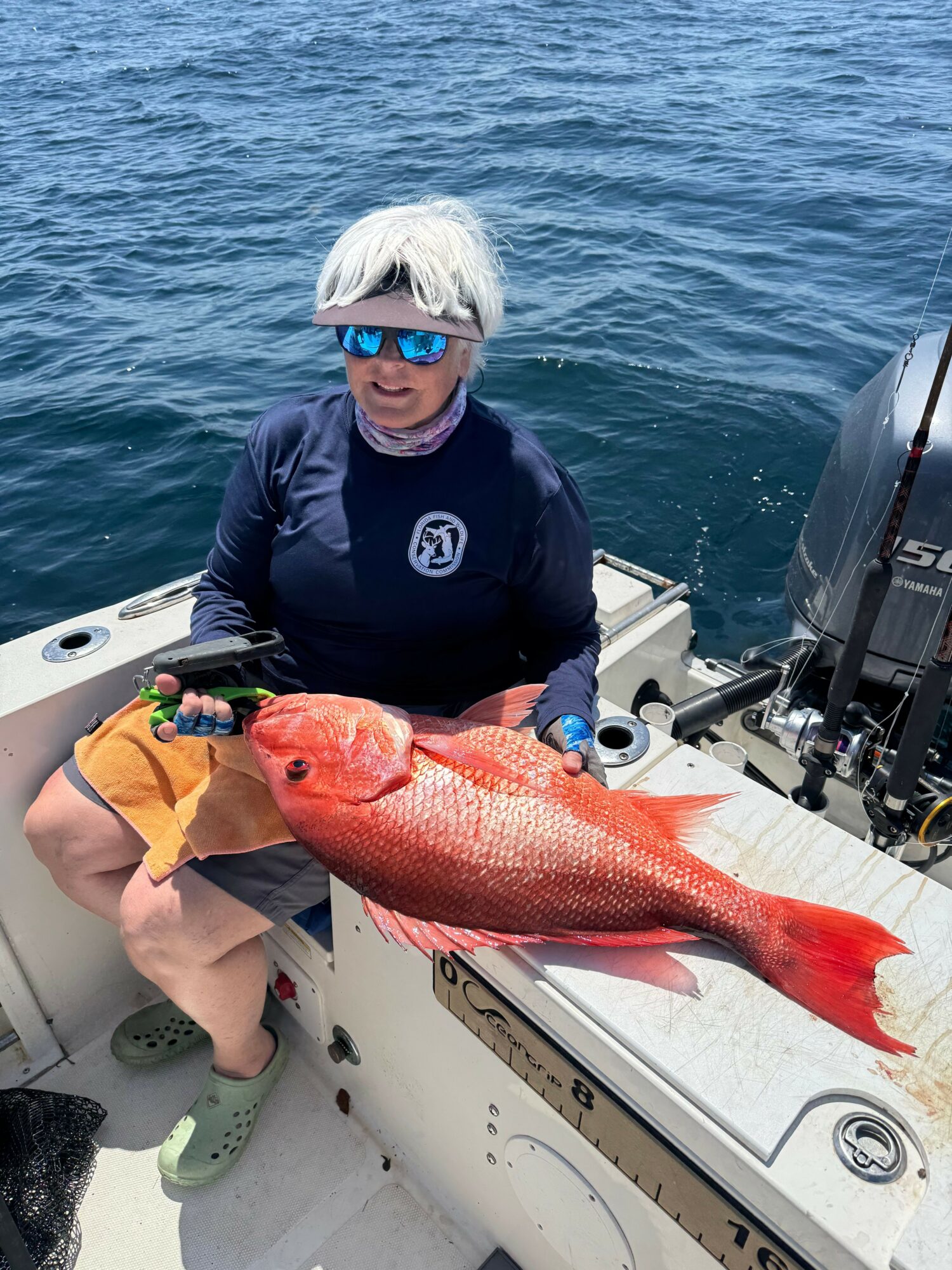 Person with white hair and sunglasses holding a large red fish on a boat.