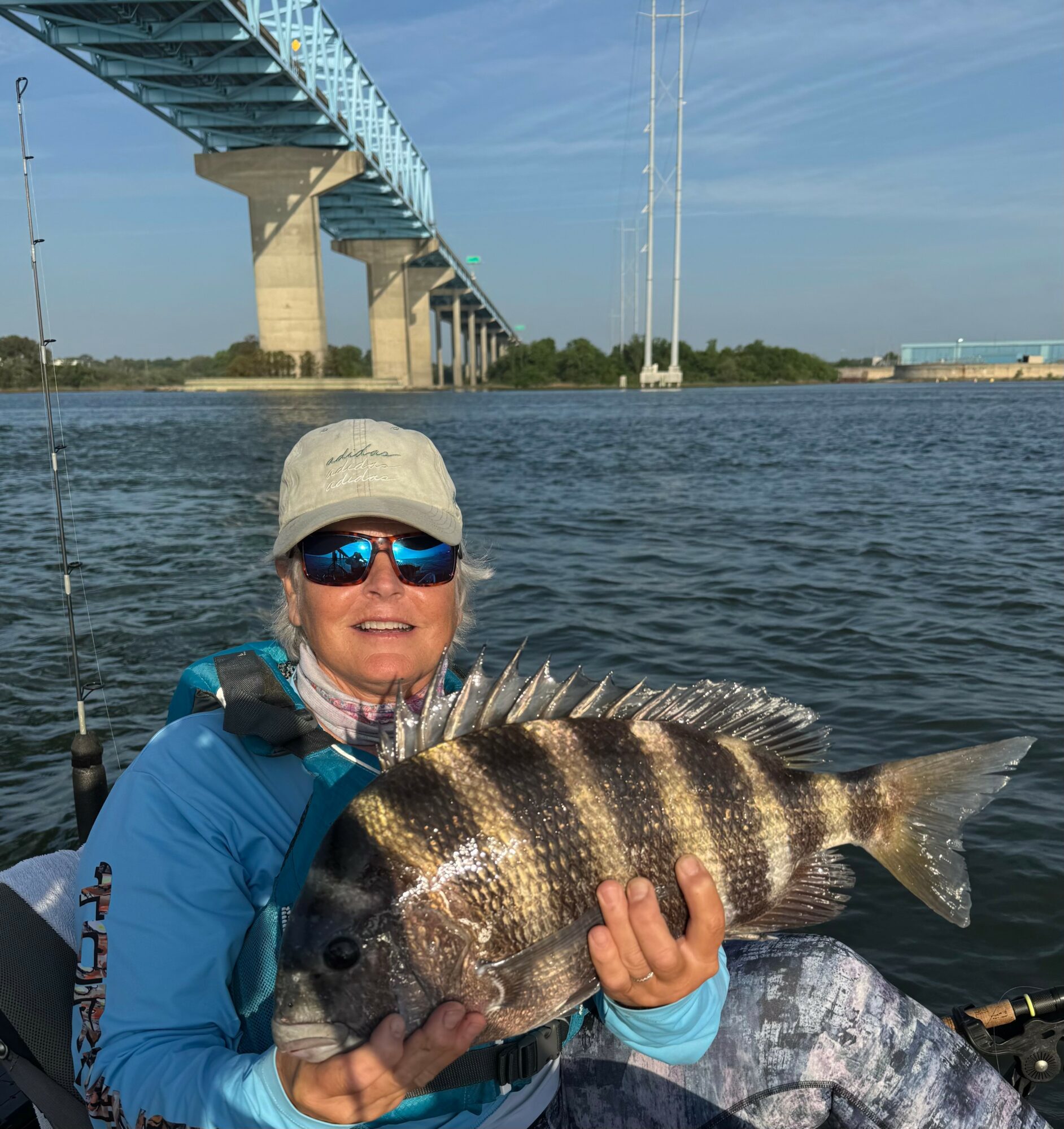 Person in blue jacket and sunglasses holding a large fish with a bridge and water in background.