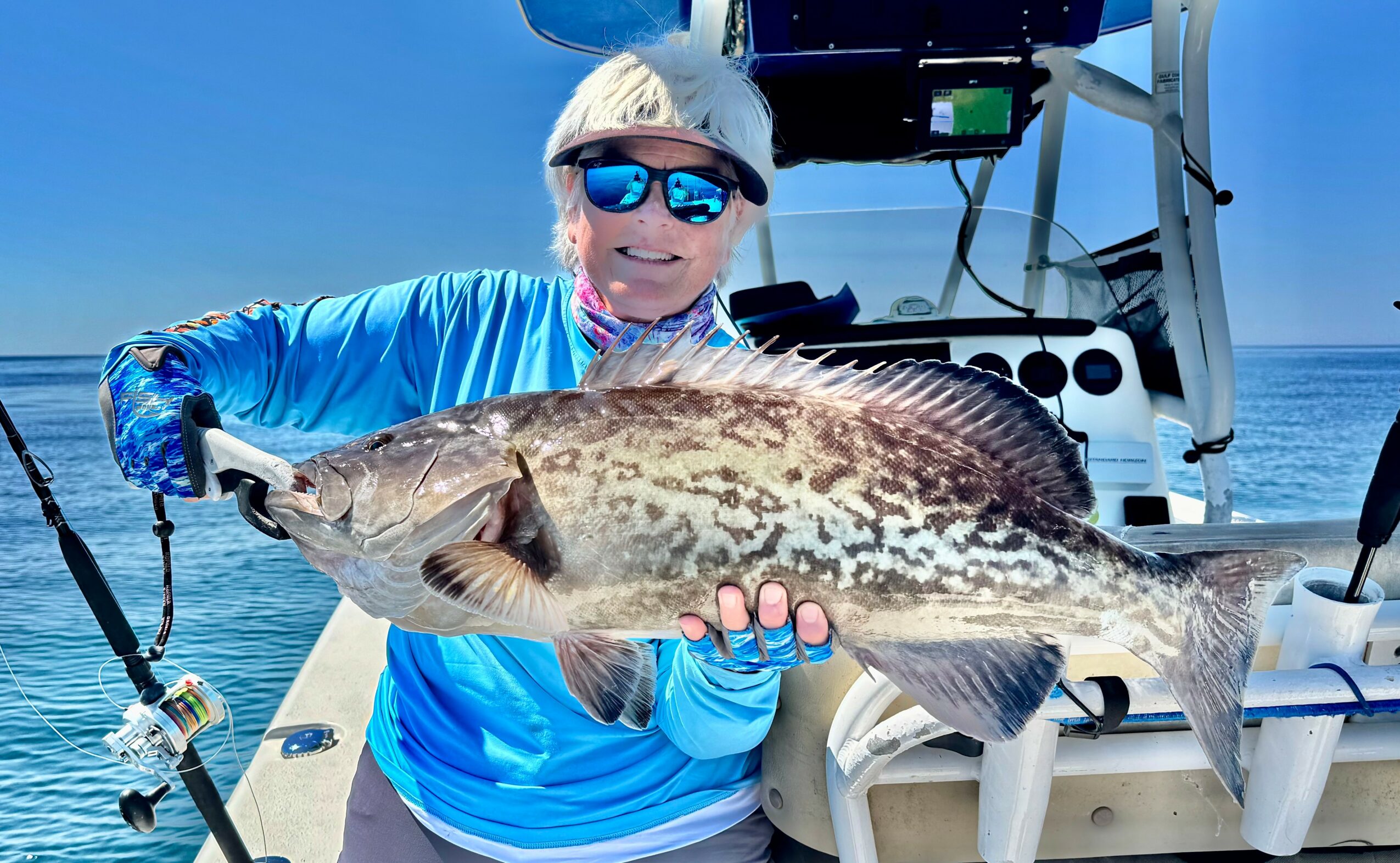 Person wearing sunglasses and a blue jacket holding a large fish on a boat, ocean in background.