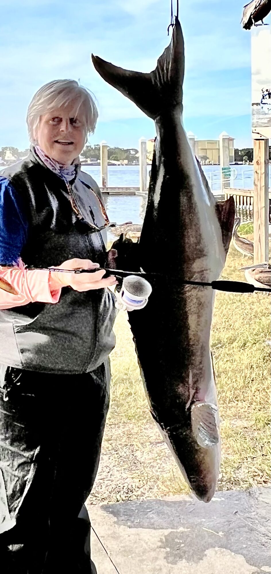Person holding a large fish outdoors near water with buildings in background.