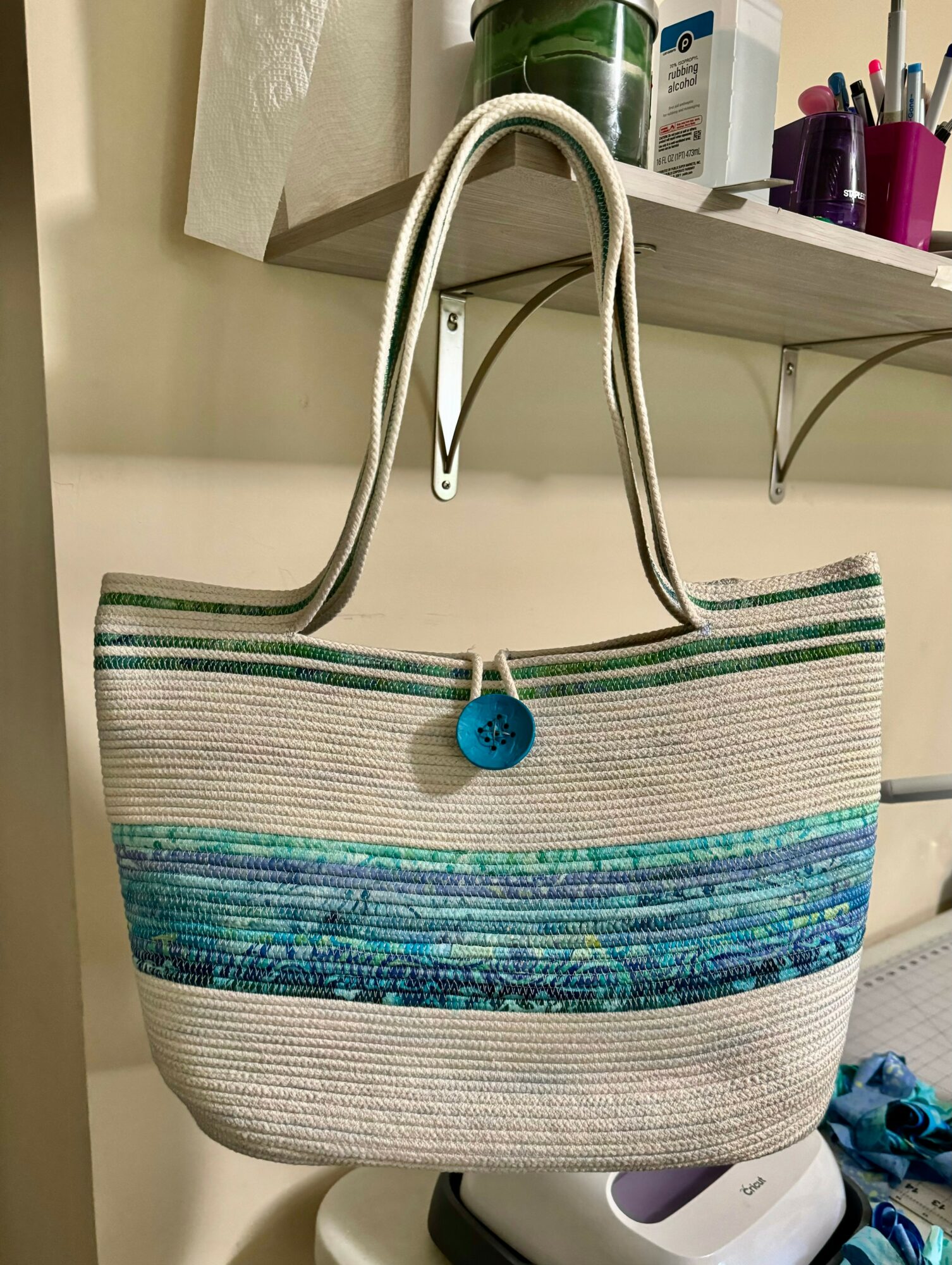 Striped tote bag with blue, green, and beige horizontal lines hanging on a wall, with a blue button closure.