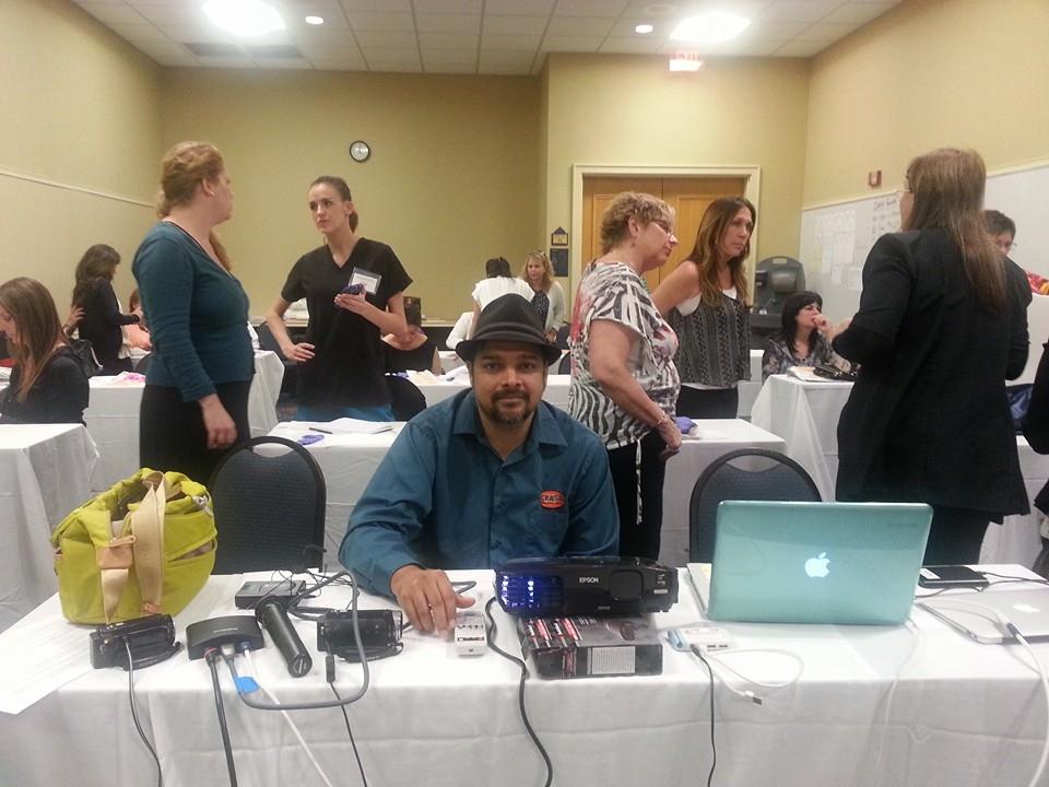 Man sitting at a table with electronic equipment, surrounded by people in a conference room.