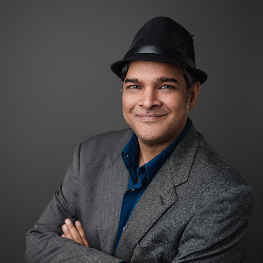Man in gray suit and black hat smiling with arms crossed against gray background.